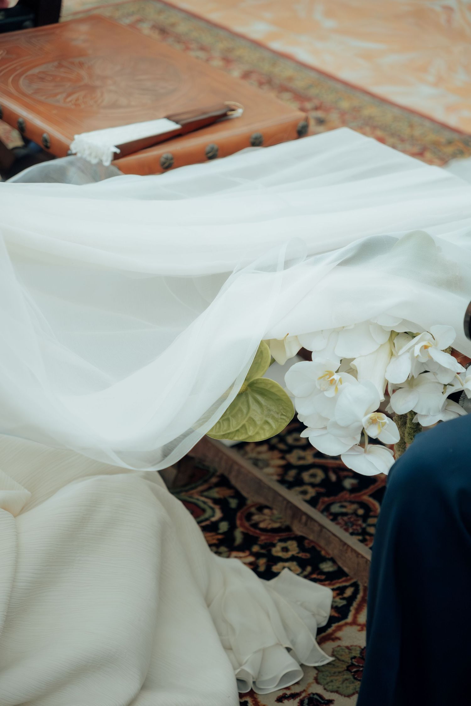 Close-up of a flowing white wedding dress draped over a chair with white orchid bouquet on a patterned carpet.