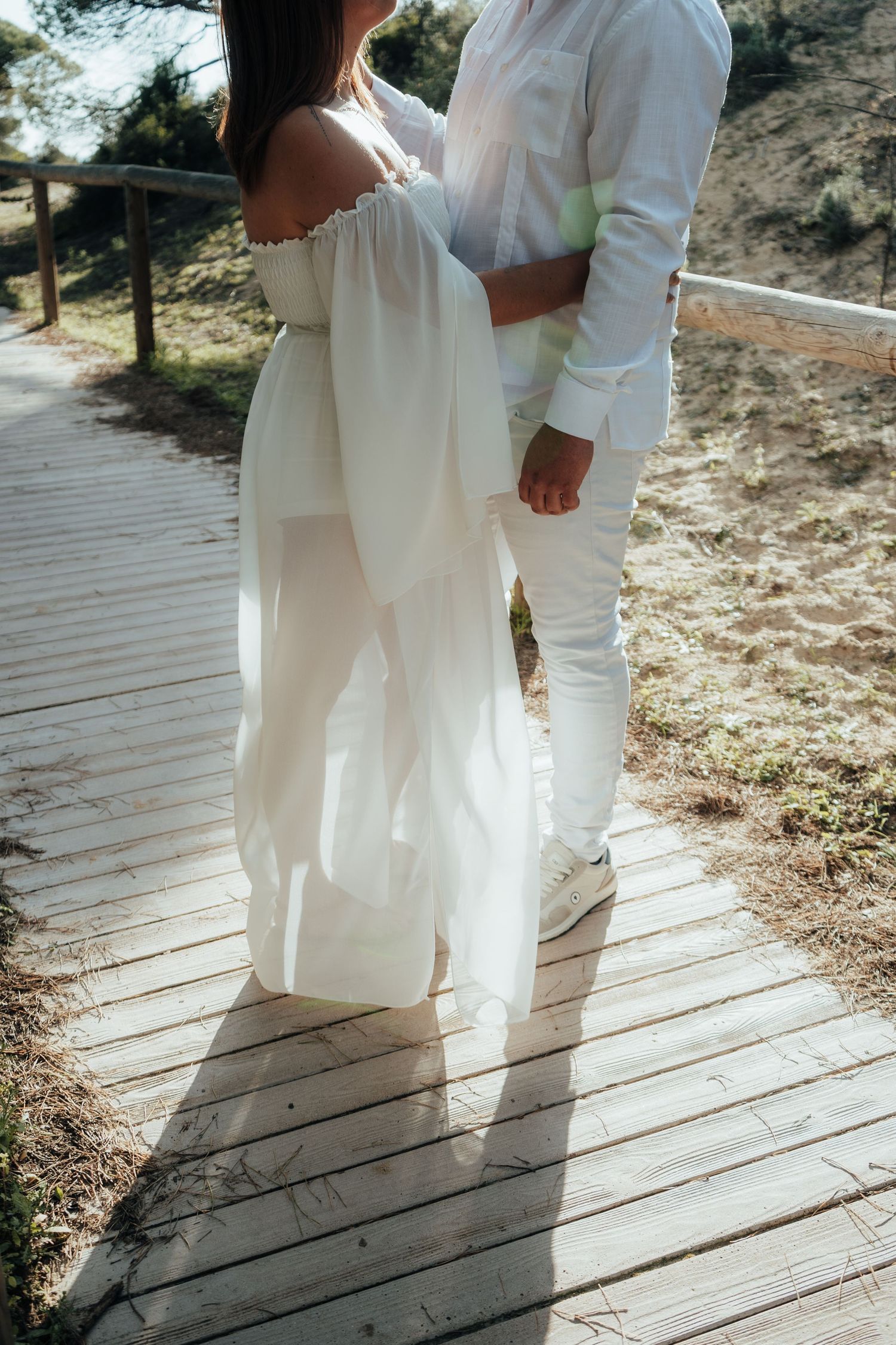 Couple in white outfits embracing on a wooden boardwalk path, woman in flowing off-shoulder dress, sunny outdoor setting.