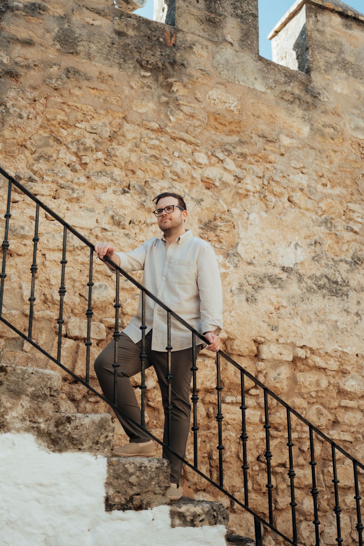 Man in glasses and linen shirt ascending stone stairs beside rustic medieval wall.