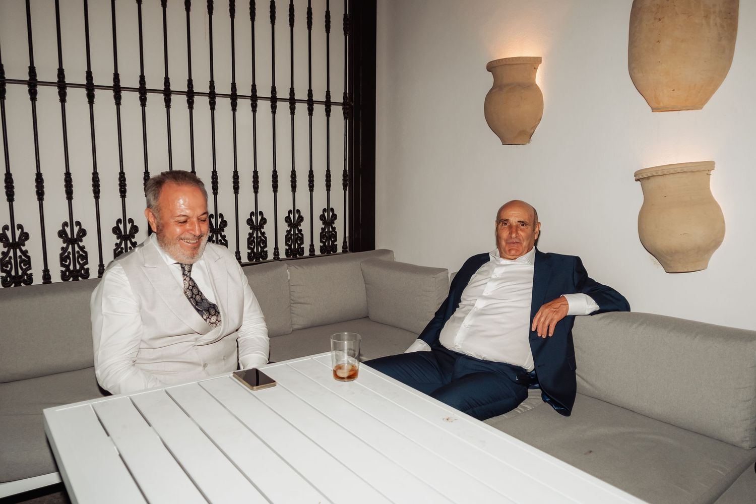 Two men relaxing on white sofas in an elegant lounge with decorative clay pots on the wall.
