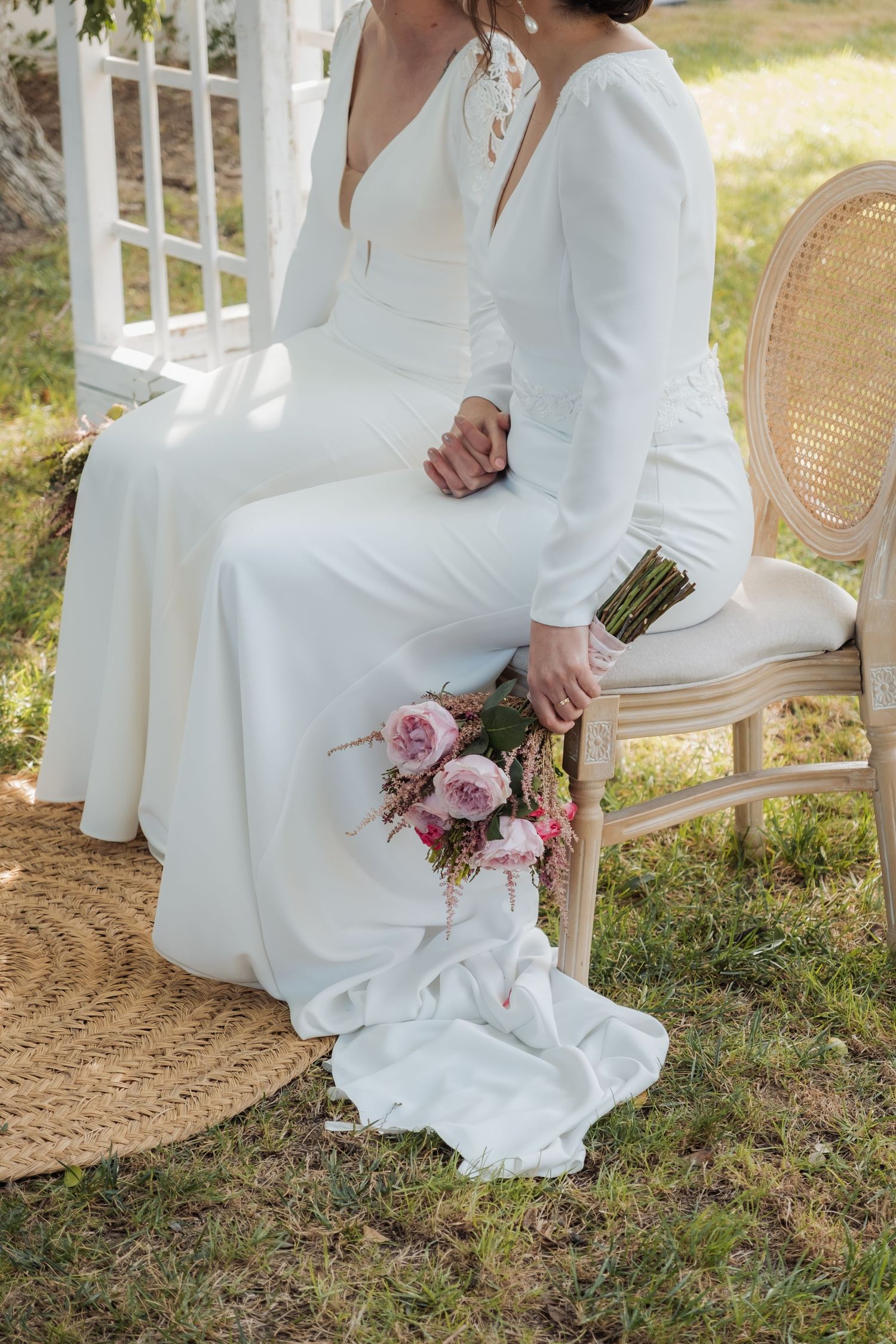 A bride in a white long-sleeve gown holds a pink floral bouquet while seated on a rattan chair outdoors.