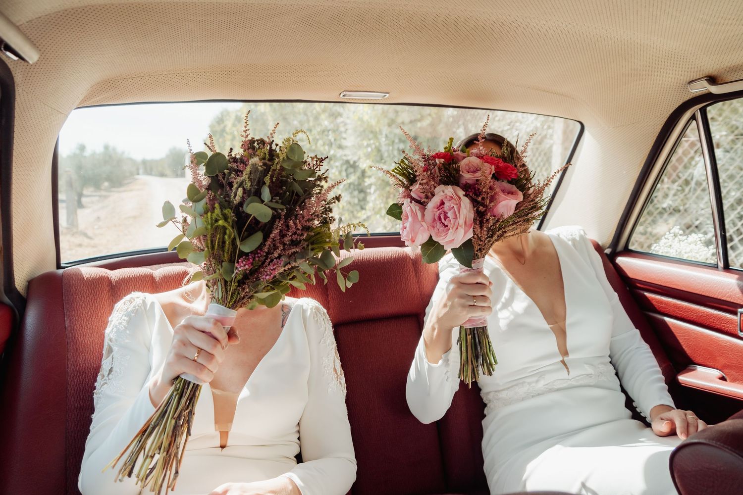 Two brides hide their faces behind wedding bouquets while sitting in a vintage red car interior.