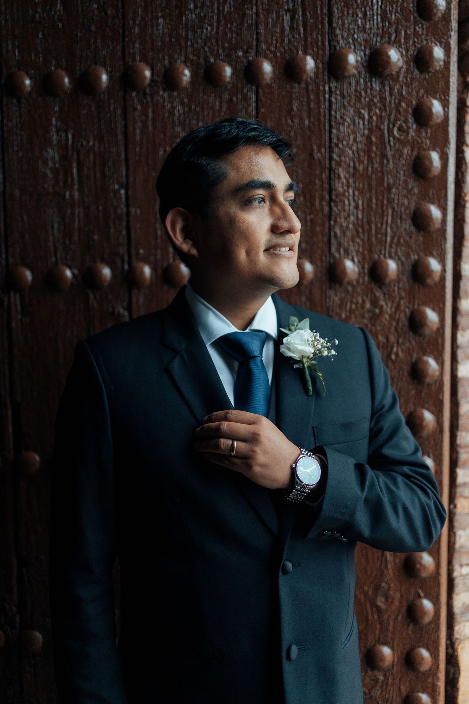Handsome groom in a dark suit with boutonniere and watch standing confidently by a rustic wooden door.