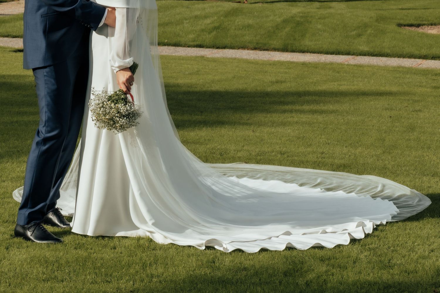 Bride and groom stand on green grass, showcasing the stunning cathedral-length train of her white wedding gown.