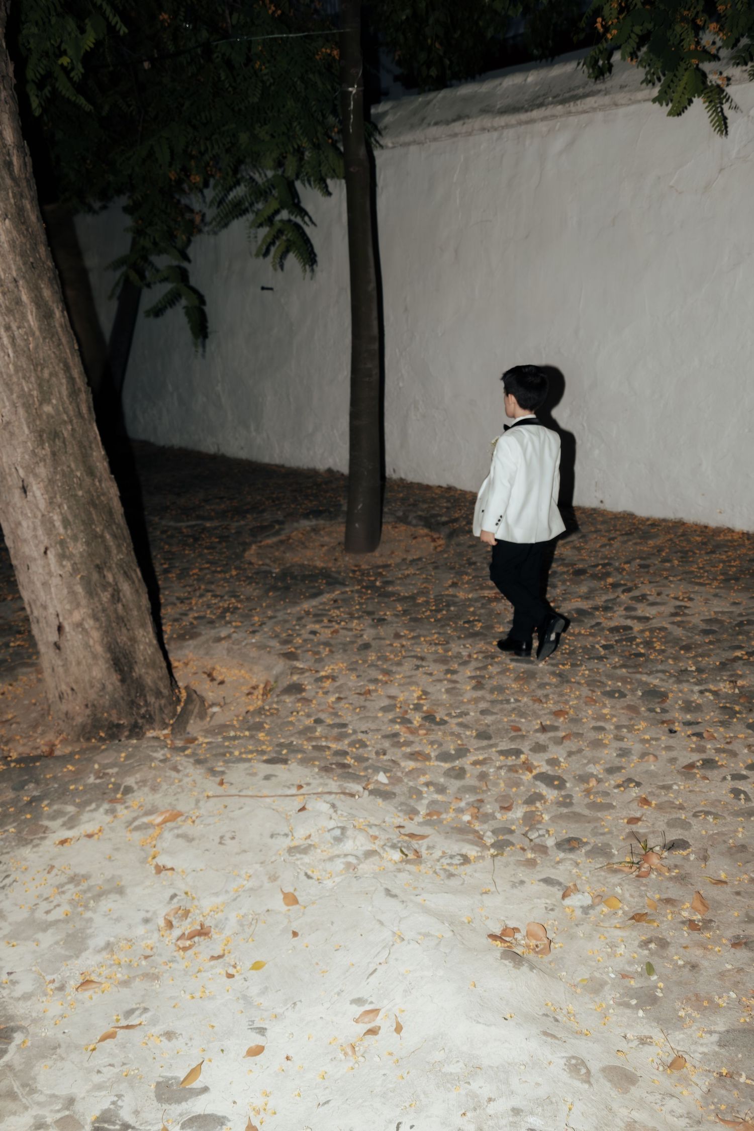 Young boy in formal white tuxedo walking alone in courtyard at night.