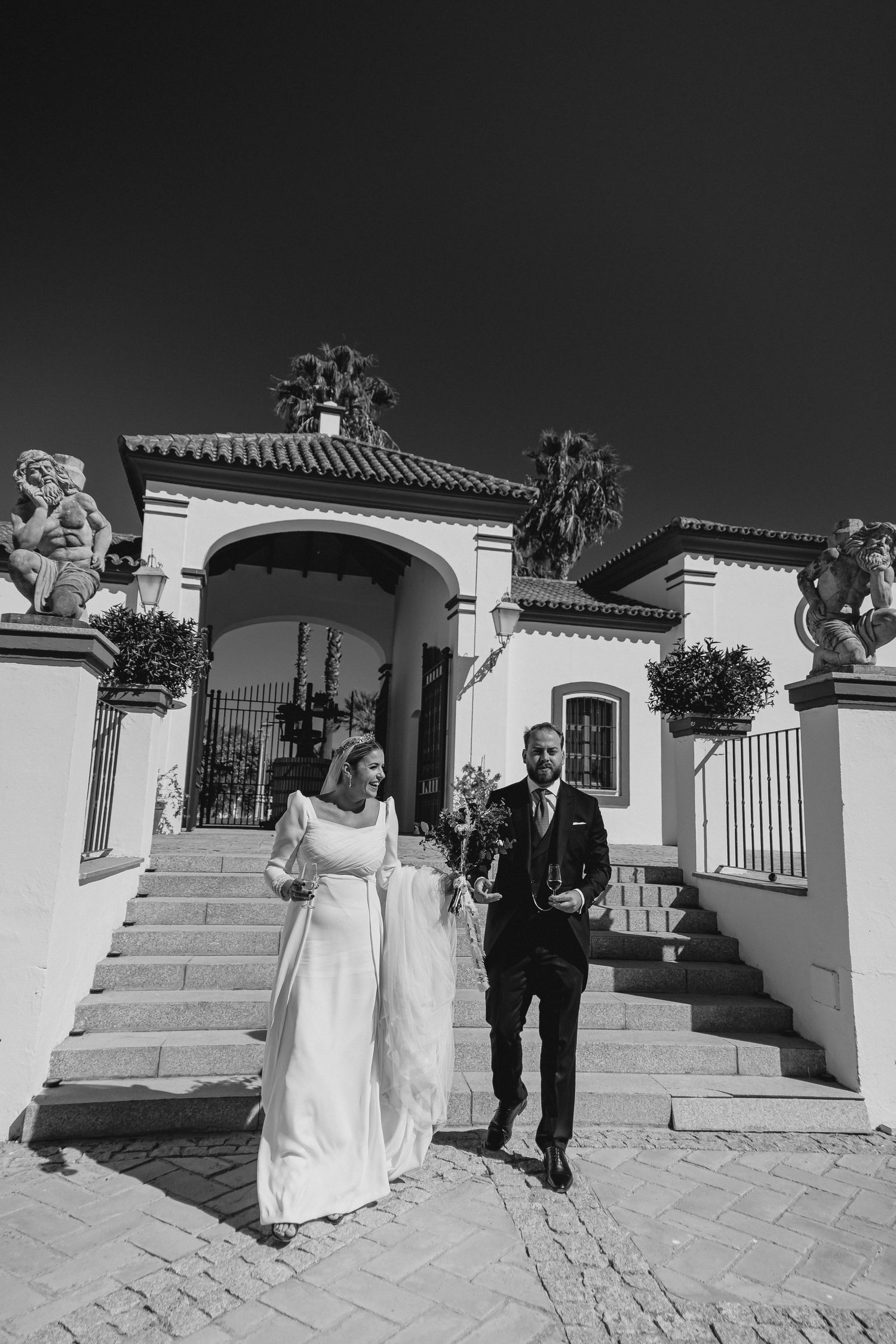 Black and white photo of bride and groom walking from ornate Spanish estate entrance with statues.