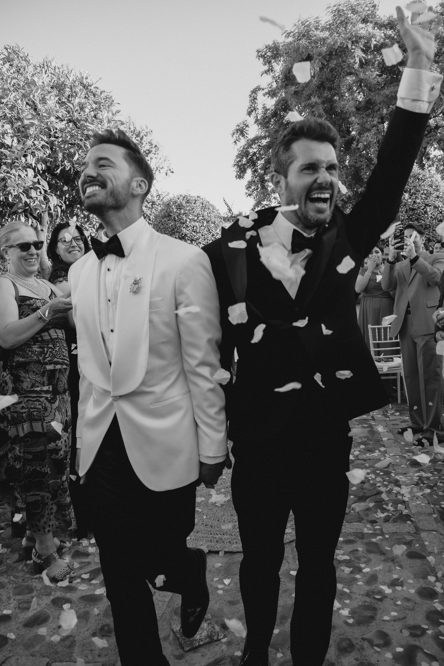 Black and white photo of two joyful grooms walking through confetti after outdoor wedding ceremony.