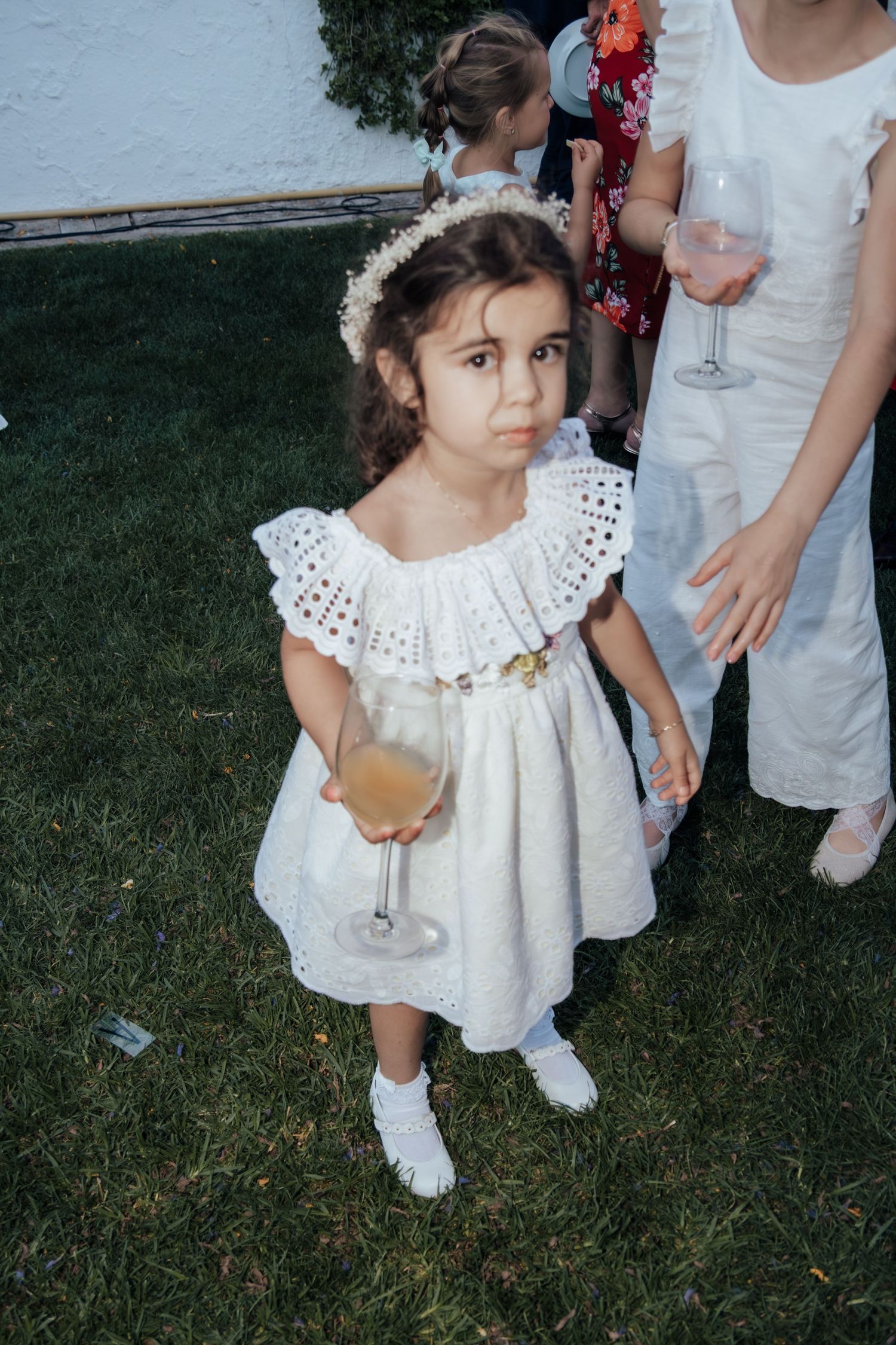 Young flower girl in white dress holding champagne glass at outdoor wedding.