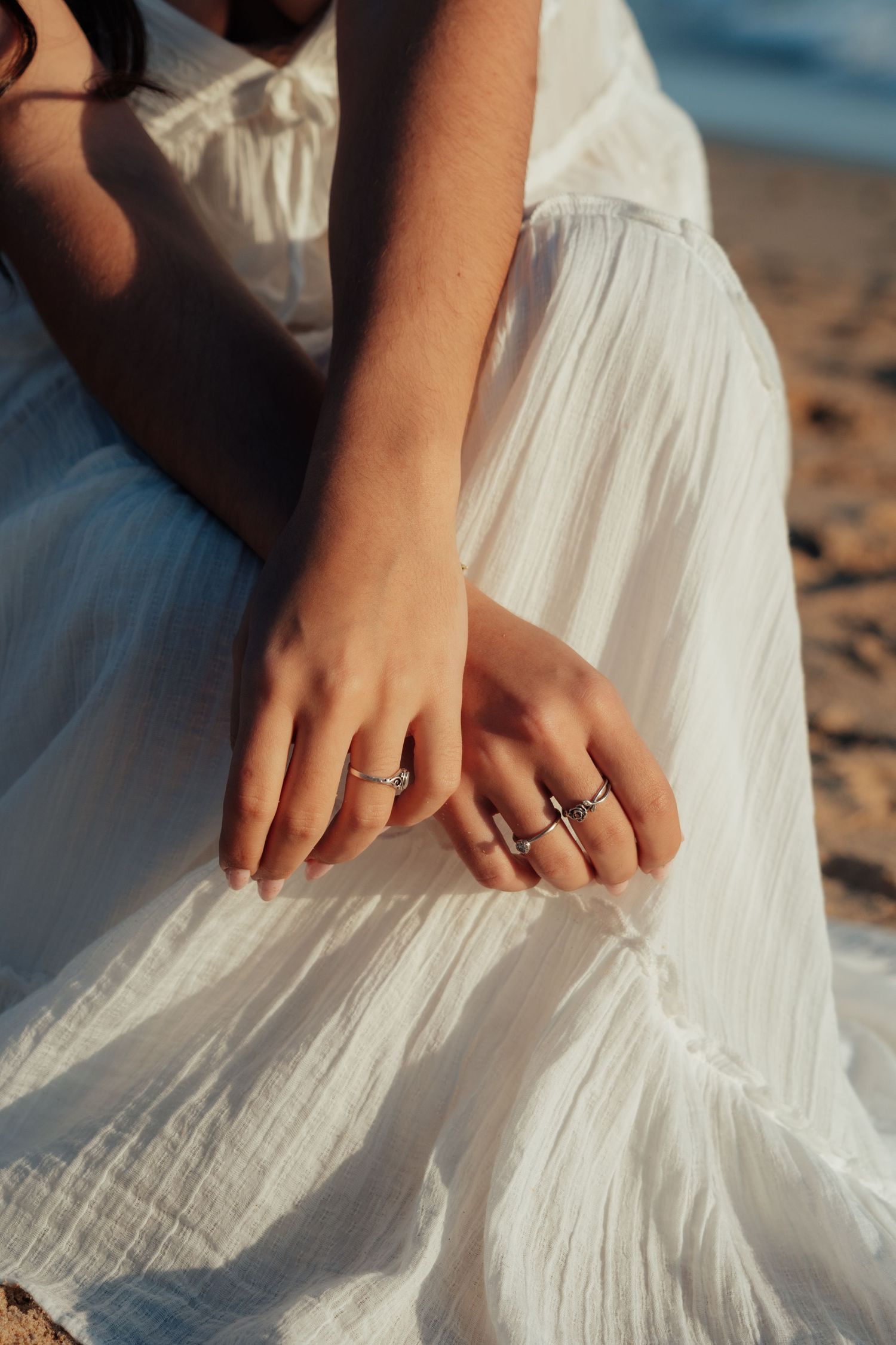 Close-up of two hands adorned with silver rings resting on a white flowing dress at a beach.