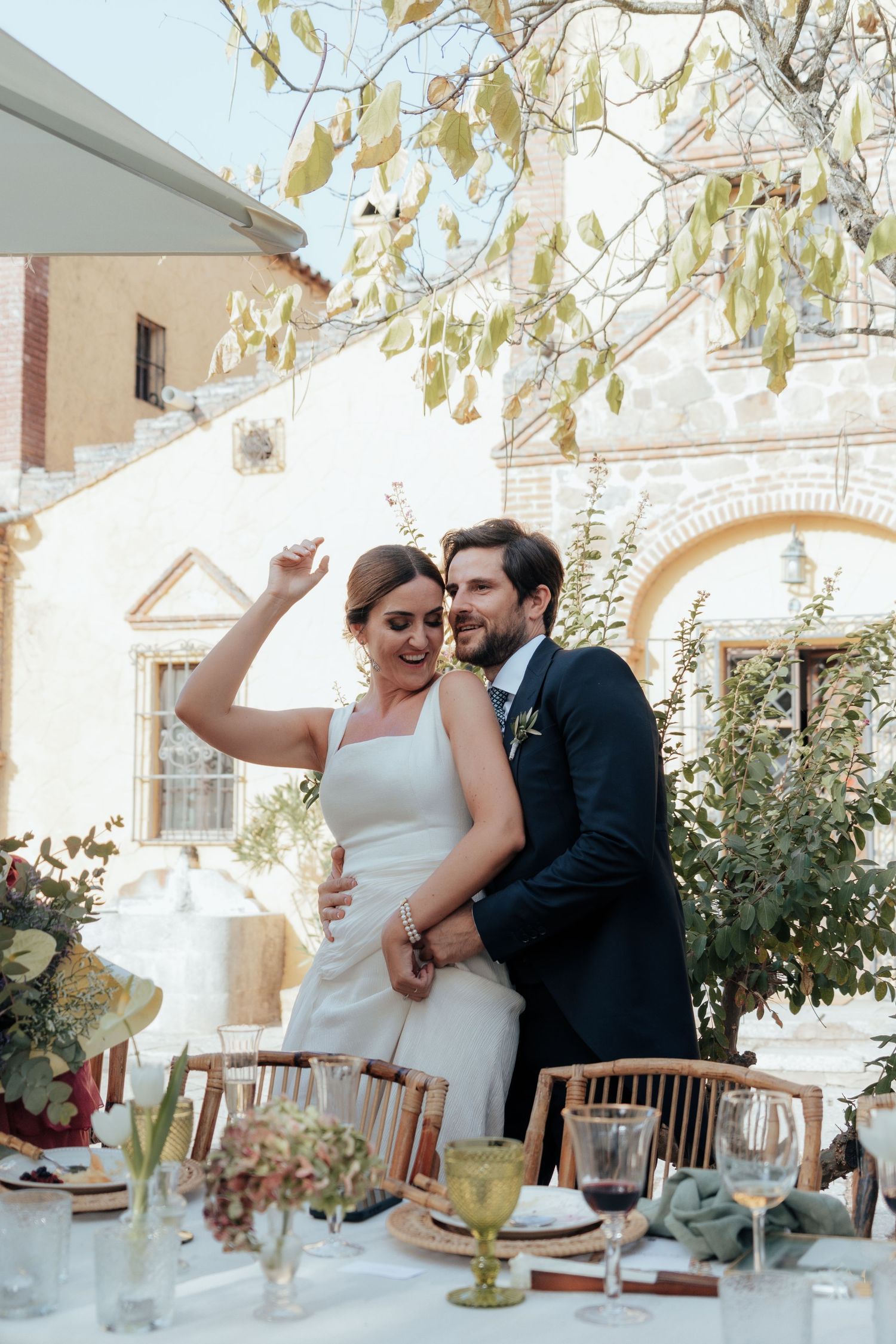 Bride and groom embracing at elegant outdoor wedding reception table with stone building backdrop.