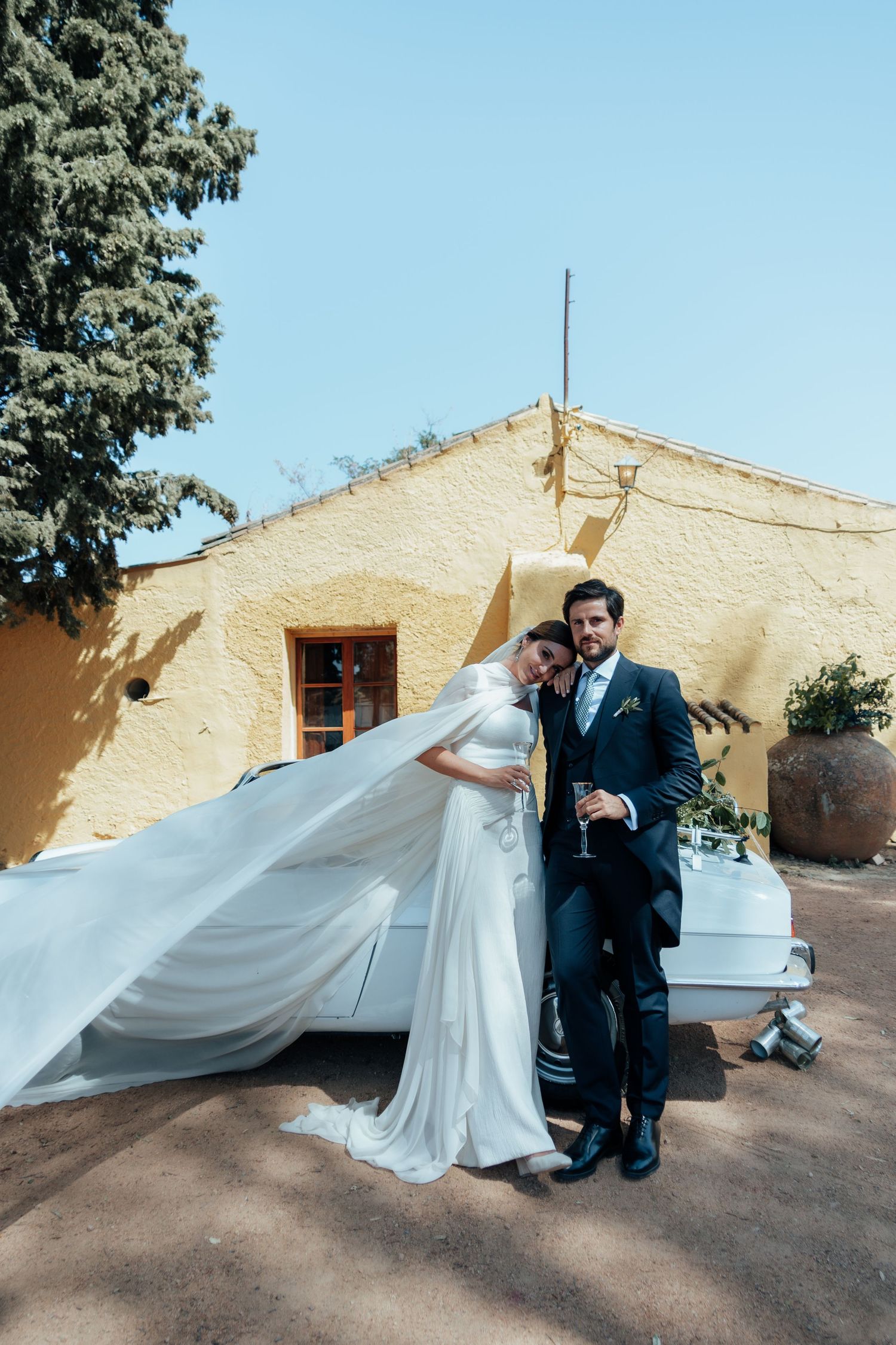 Elegant bride in flowing white gown with veil leaning on groom in navy suit beside vintage white car at rustic venue.