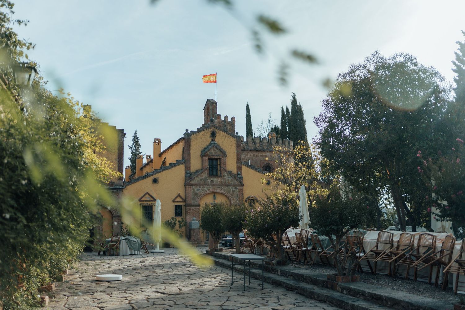 Historic yellow stone building with Spanish flag flying above surrounded by lush trees.