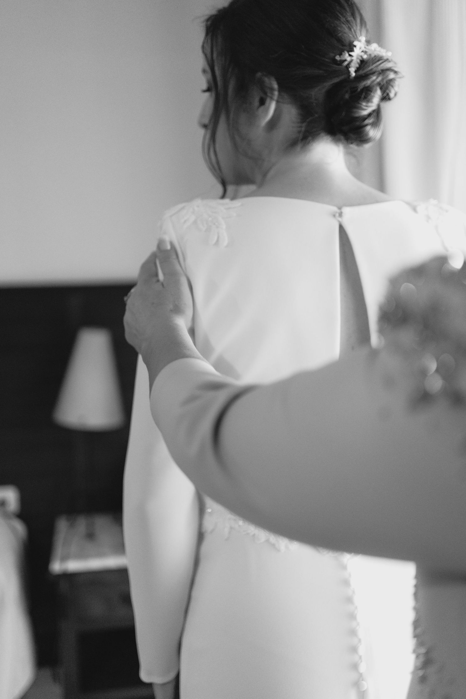 Black and white photo of bride in white gown with lace back being helped to dress.