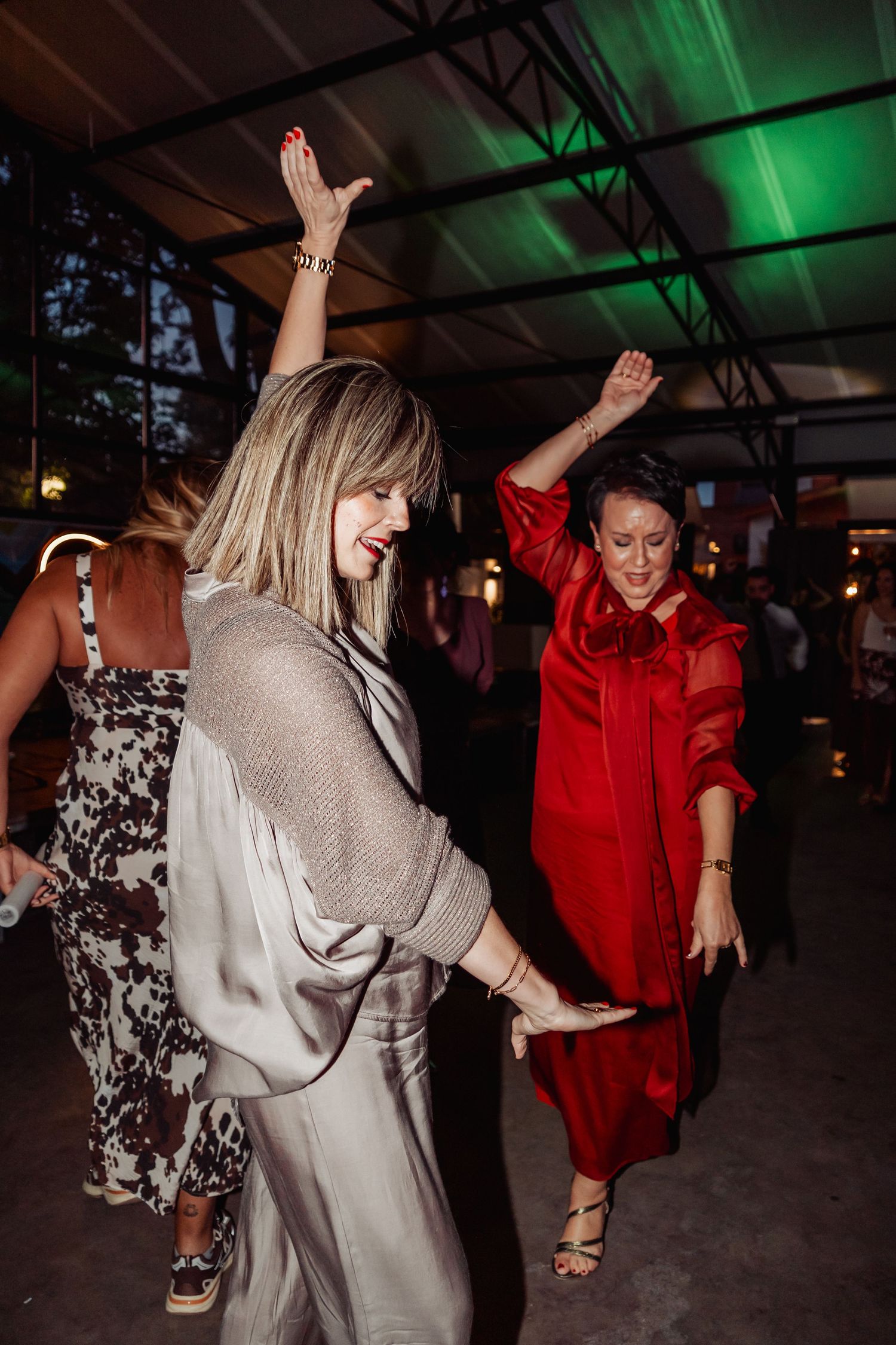 Two women dancing joyfully at an evening party, one in silver dress and one in red outfit, under green lights.