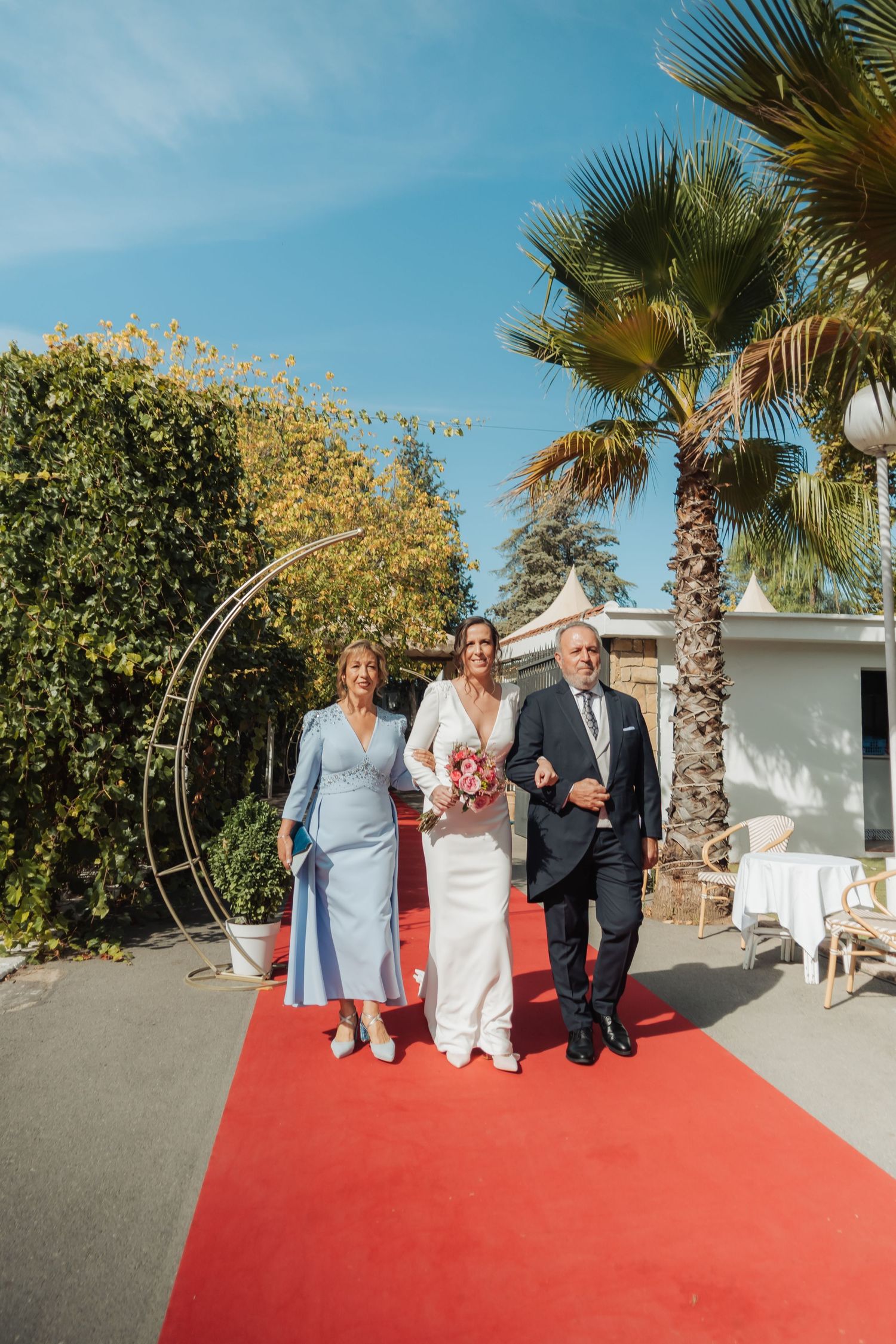 Bride in white gown walks red carpet with parents at outdoor wedding venue.