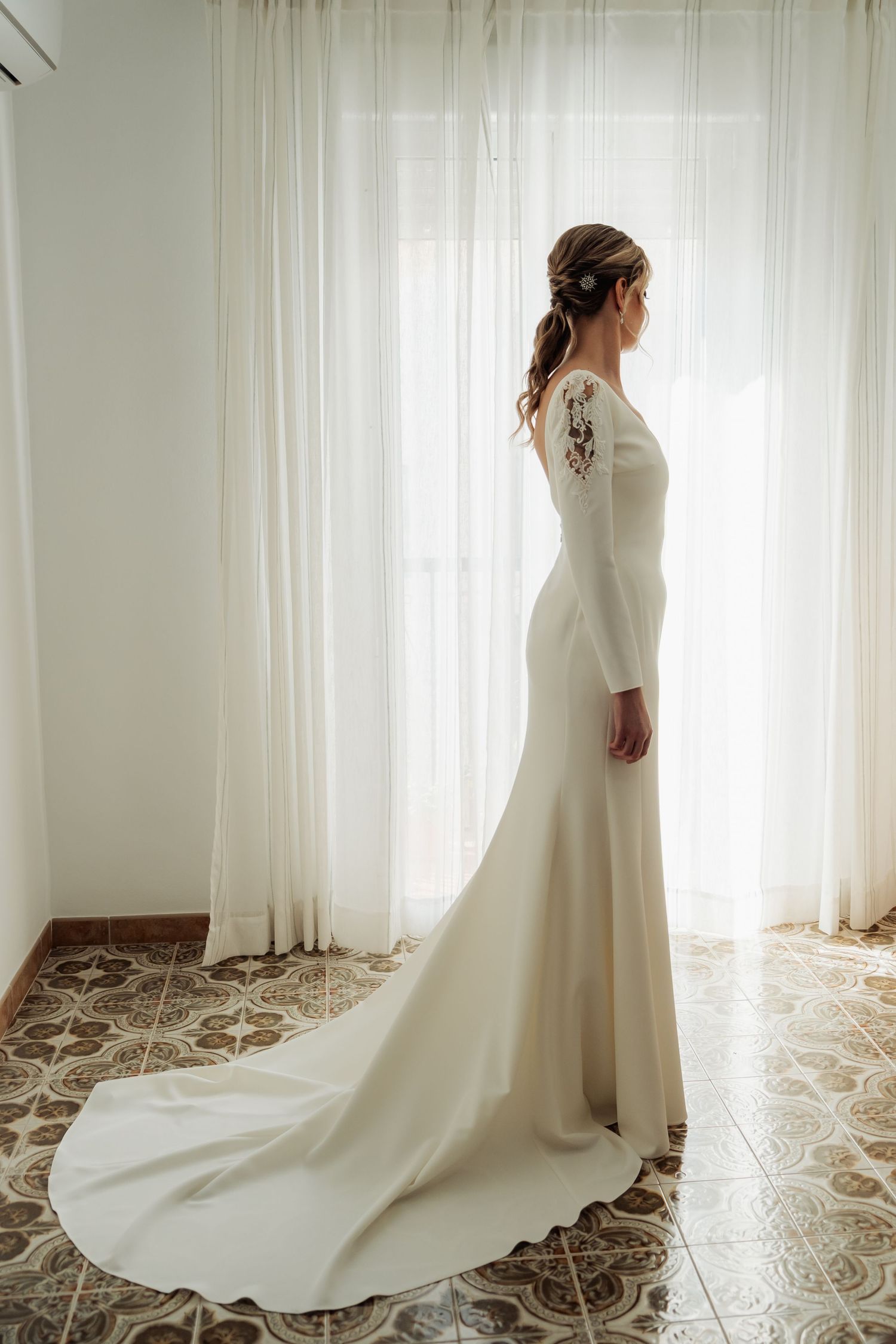 Bride in fitted long-sleeve white wedding gown with train standing by sheer curtained window.