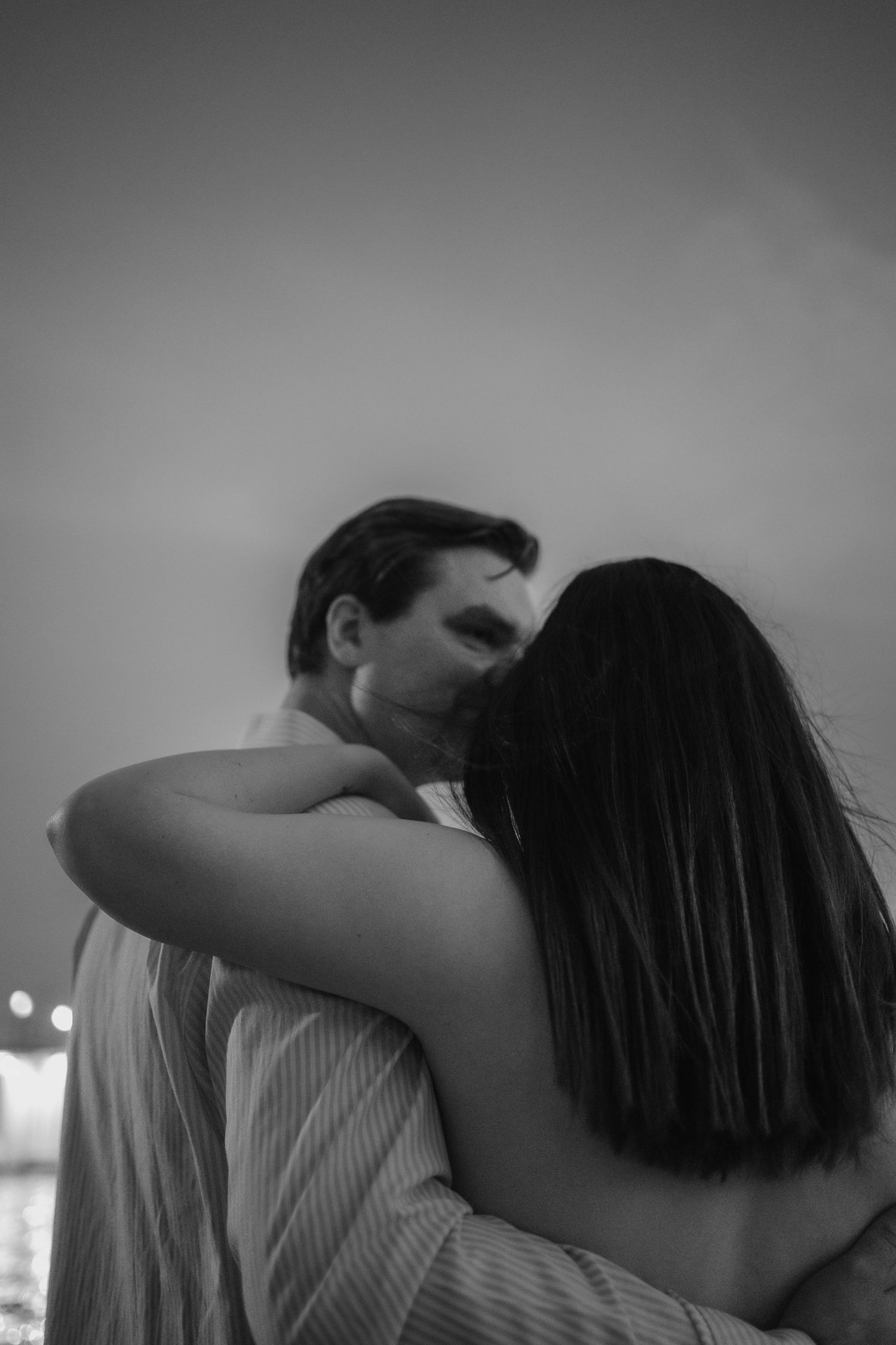 Black and white intimate portrait of a couple embracing and kissing at dusk near city lights.