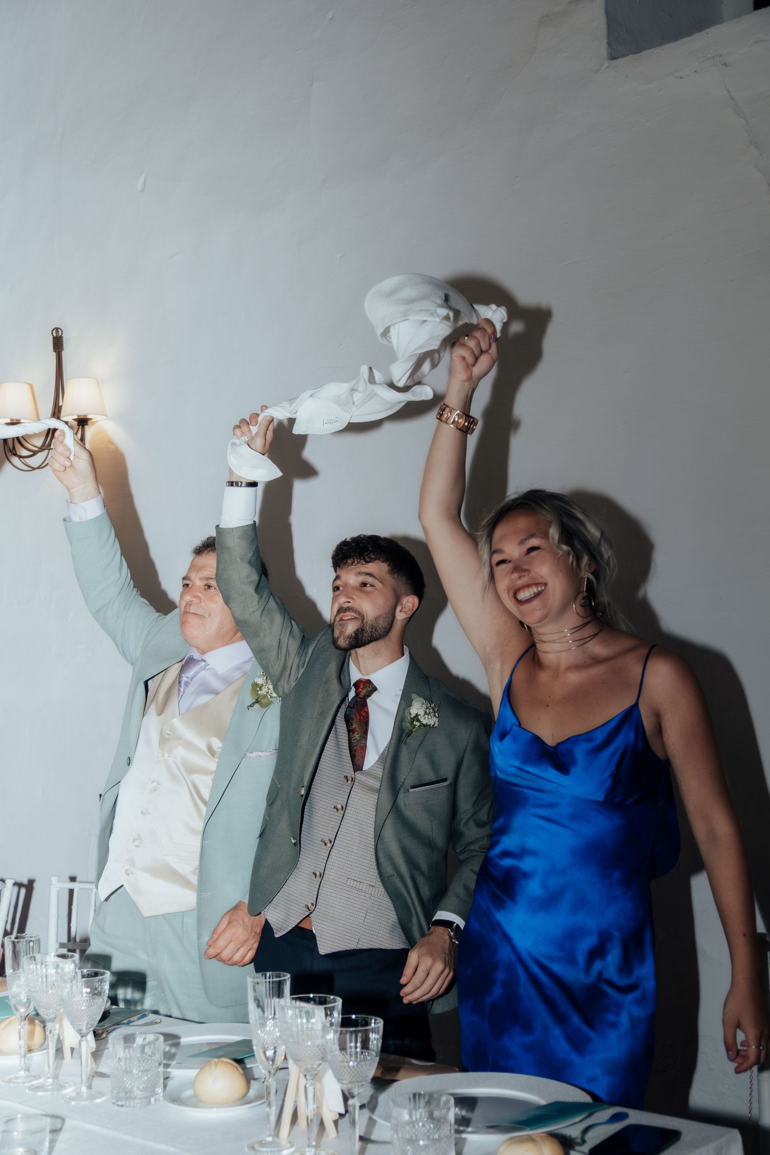 Two grooms and woman in blue dress joyfully waving white napkins at wedding reception table celebration.