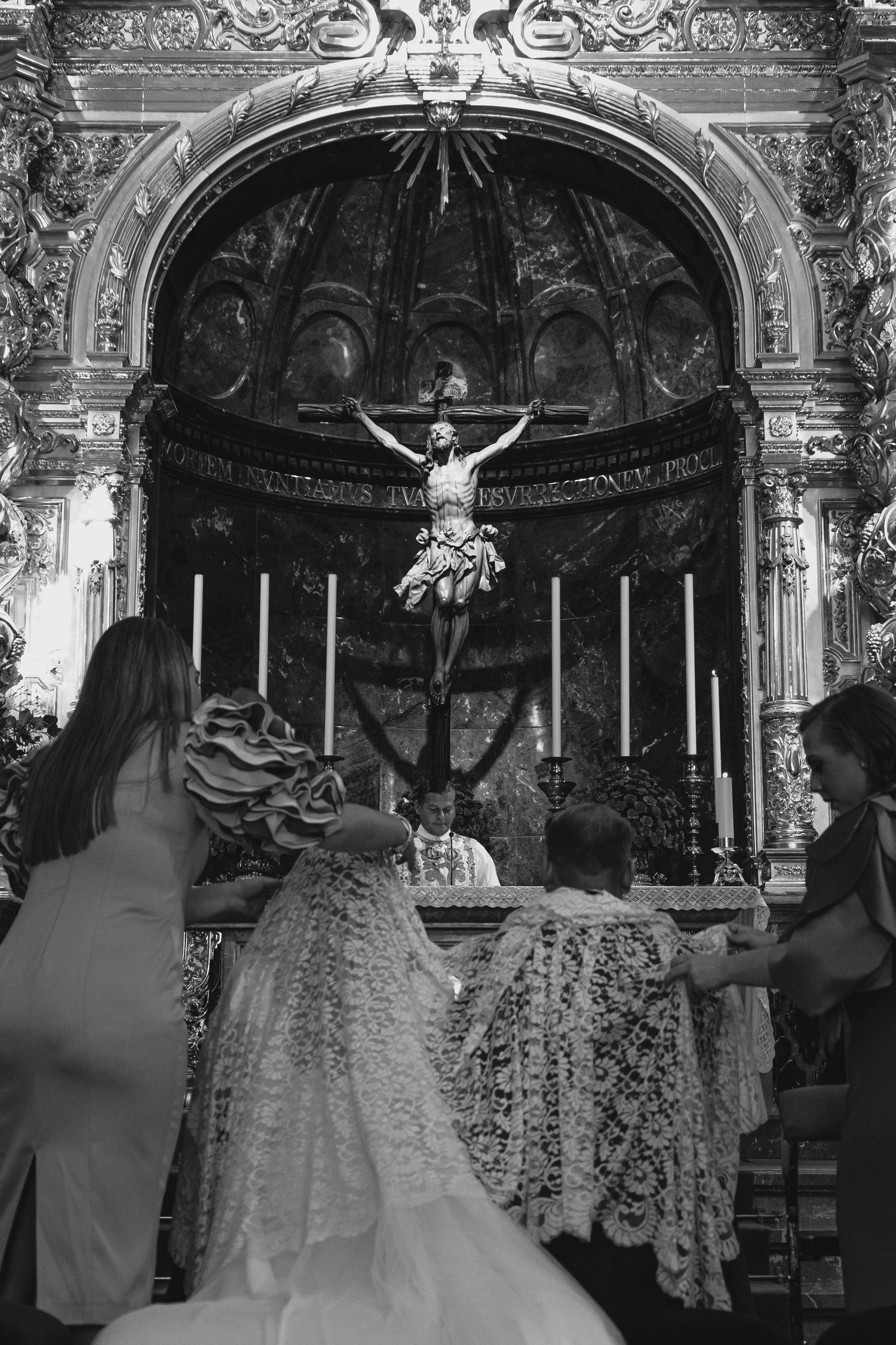 Black and white photo of bride in lace gown at baroque church altar with crucifix.
