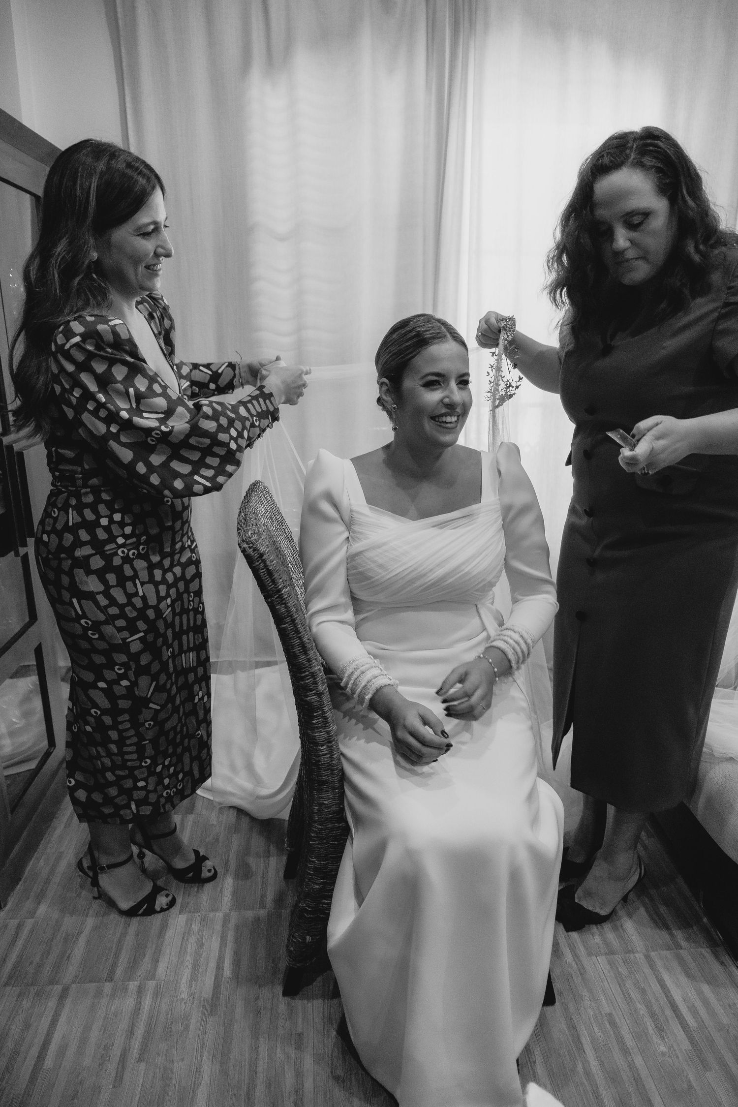 Black and white photo of smiling bride in wedding dress being assisted by two women.