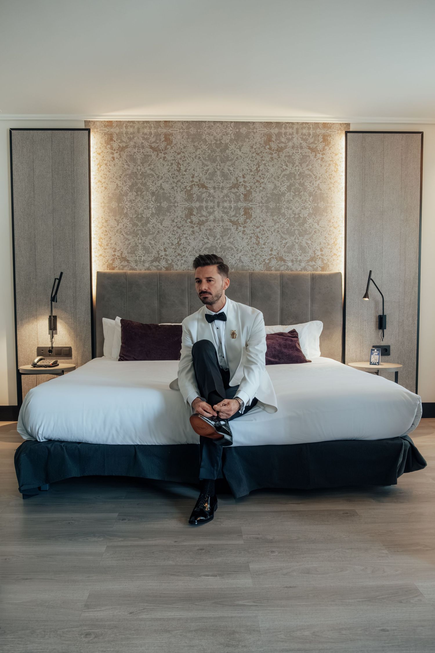 Elegant groom in white tuxedo jacket and black bow tie sits on a luxury hotel bed.