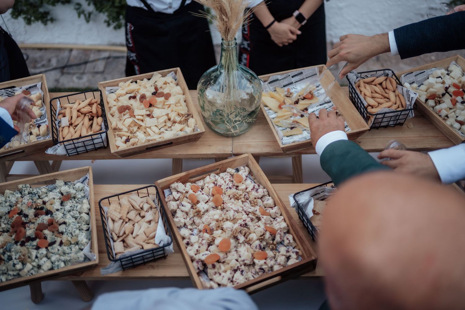 Guests selecting appetizers and snacks from wooden trays at outdoor wedding reception.