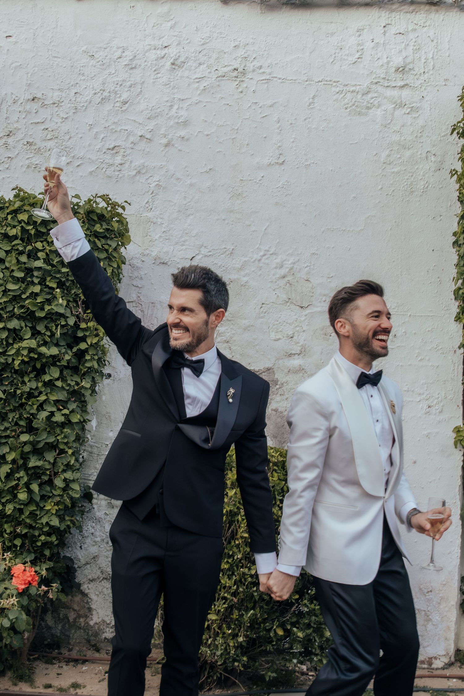 Two grooms in tuxedos celebrating joyfully, one in black and one in white jacket, holding hands at wedding.