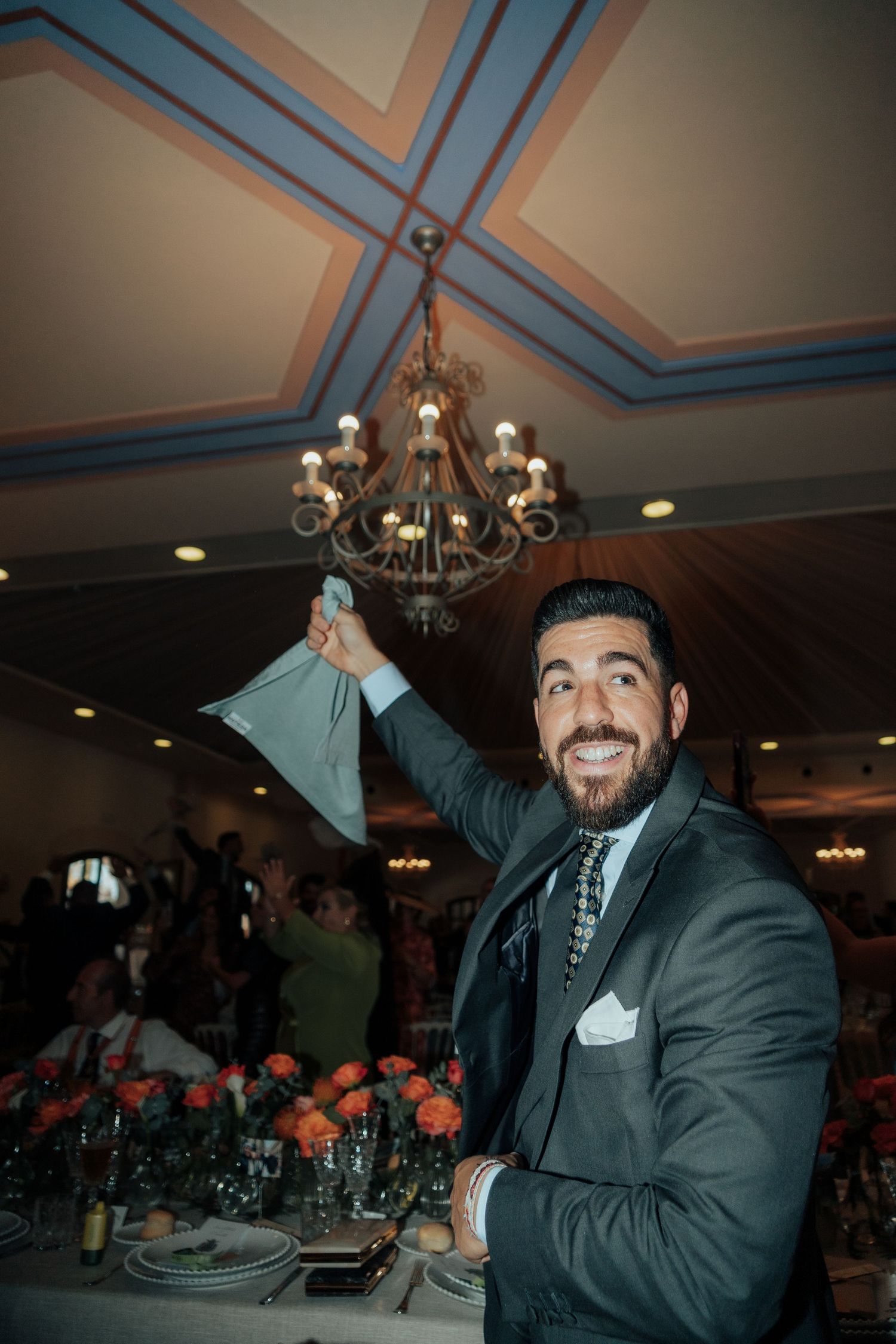 Smiling bearded man in suit raising his hand near a chandelier during a wedding reception.