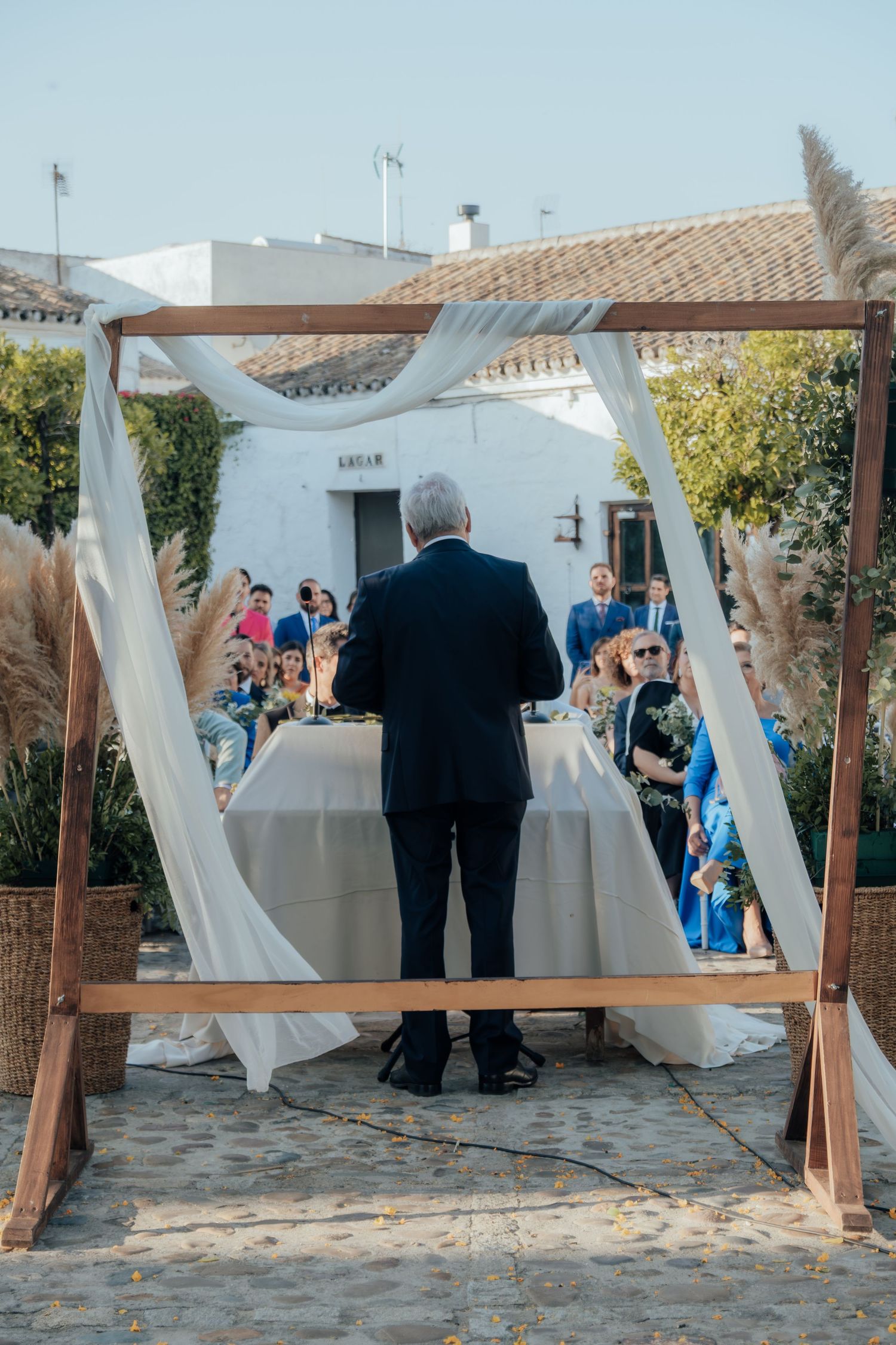 Officiant stands beneath wooden arch with white draping at outdoor wedding ceremony with seated guests.