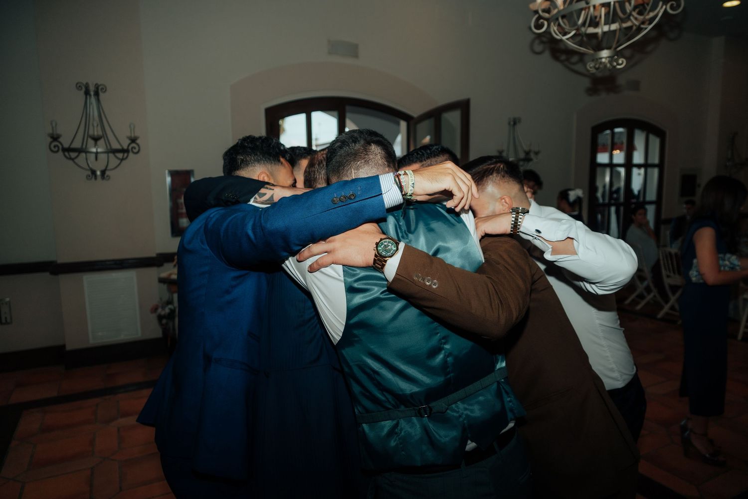 Group of men share a warm group hug celebrating together at an indoor wedding reception.