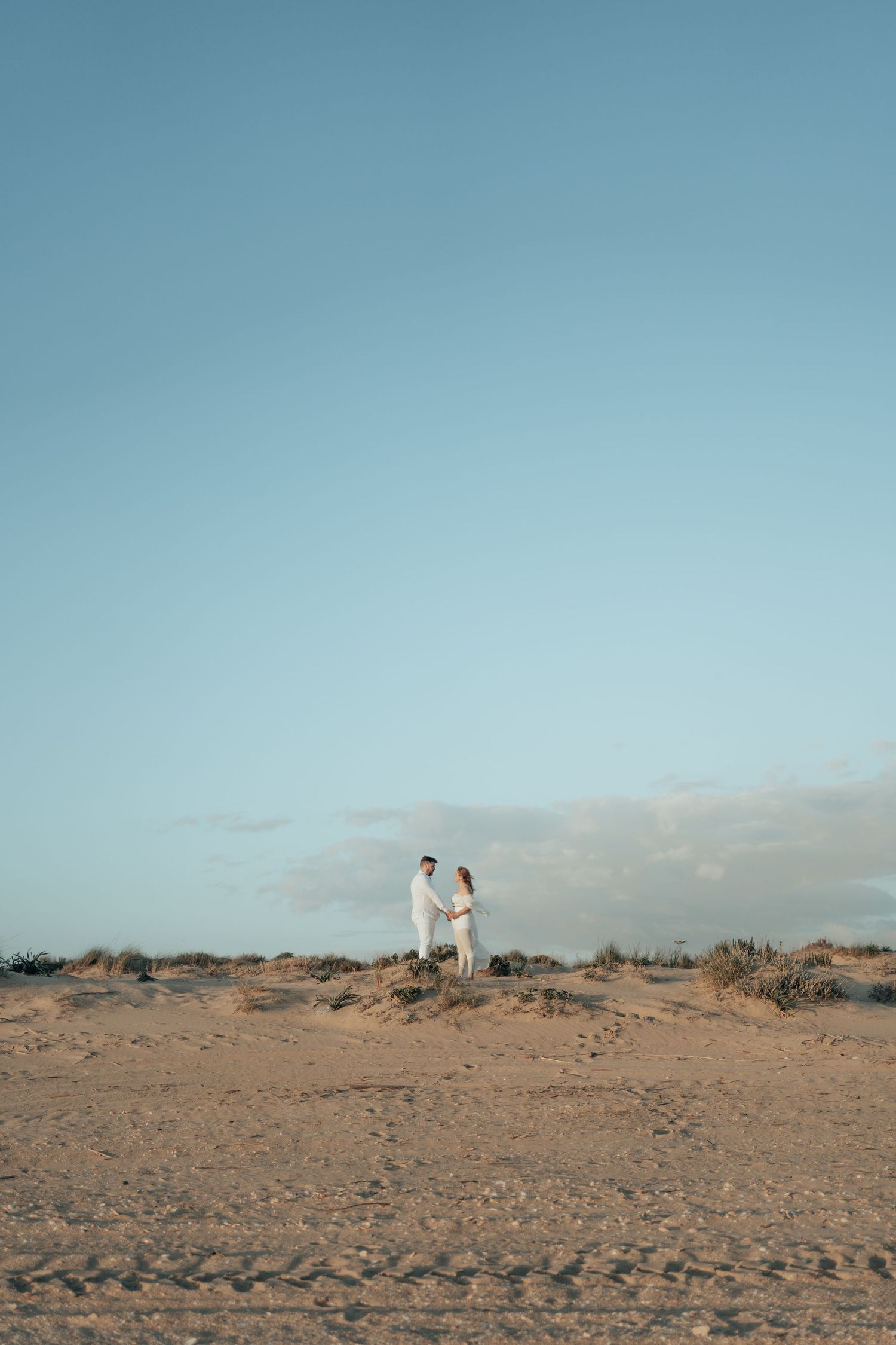 Couple in white holding hands on sandy beach dunes under a clear blue sky at golden hour.