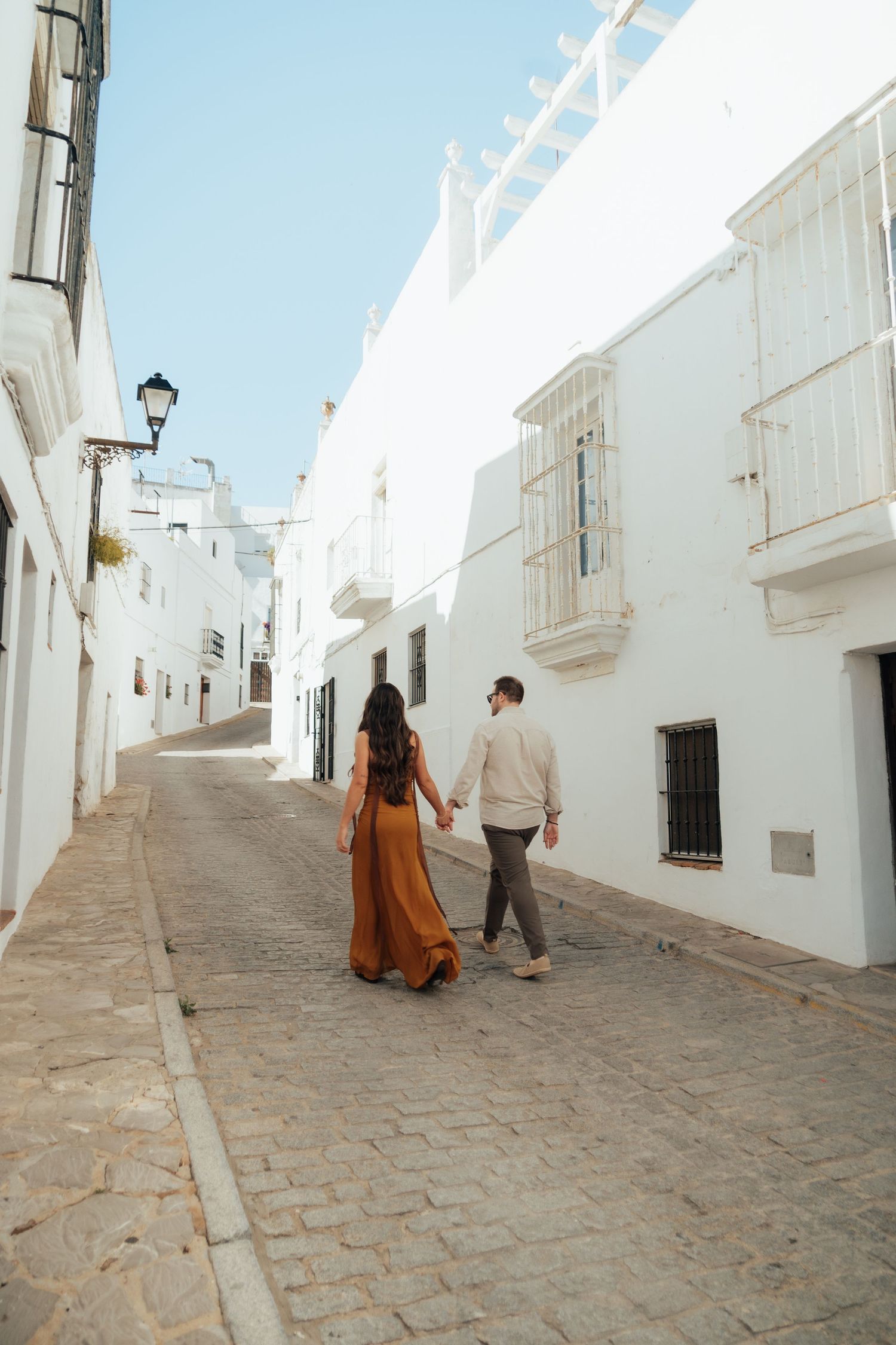 Couple holding hands walking down a charming white-washed cobblestone street in a sunny Mediterranean village.