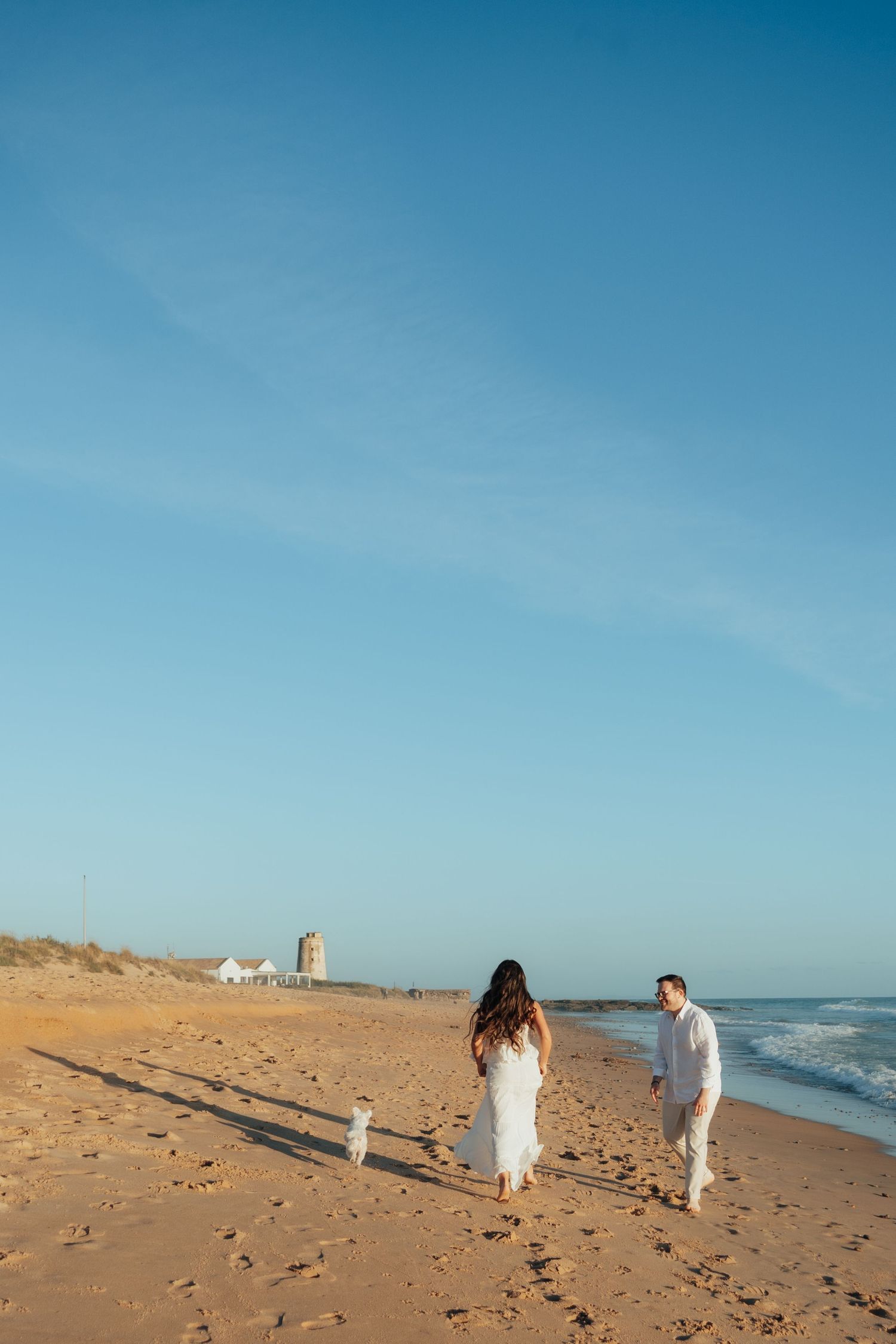 Couple in white outfits walking a small white dog on a sunny sandy beach with blue skies and ocean waves.