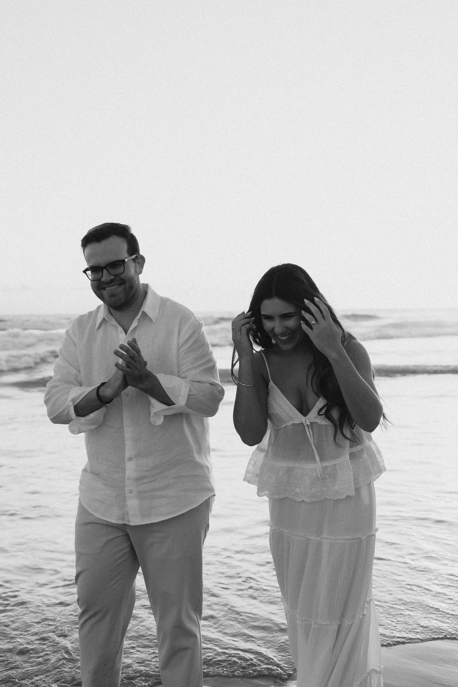 Black and white photo of a couple laughing and walking on a beach, dressed in white outfits with ocean waves behind them.
