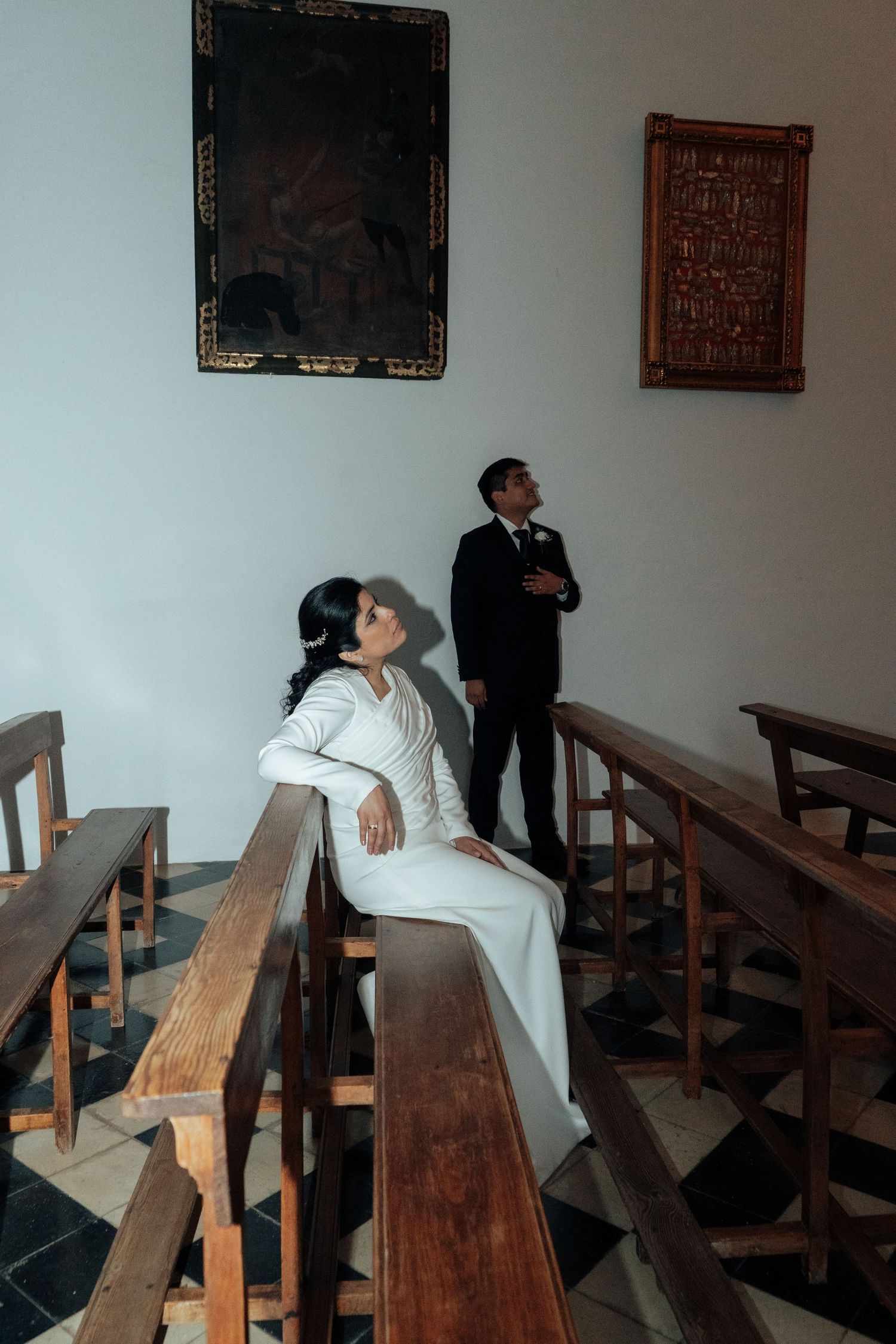 Bride in white gown sitting on wooden pew gazing upward as groom stands in church.