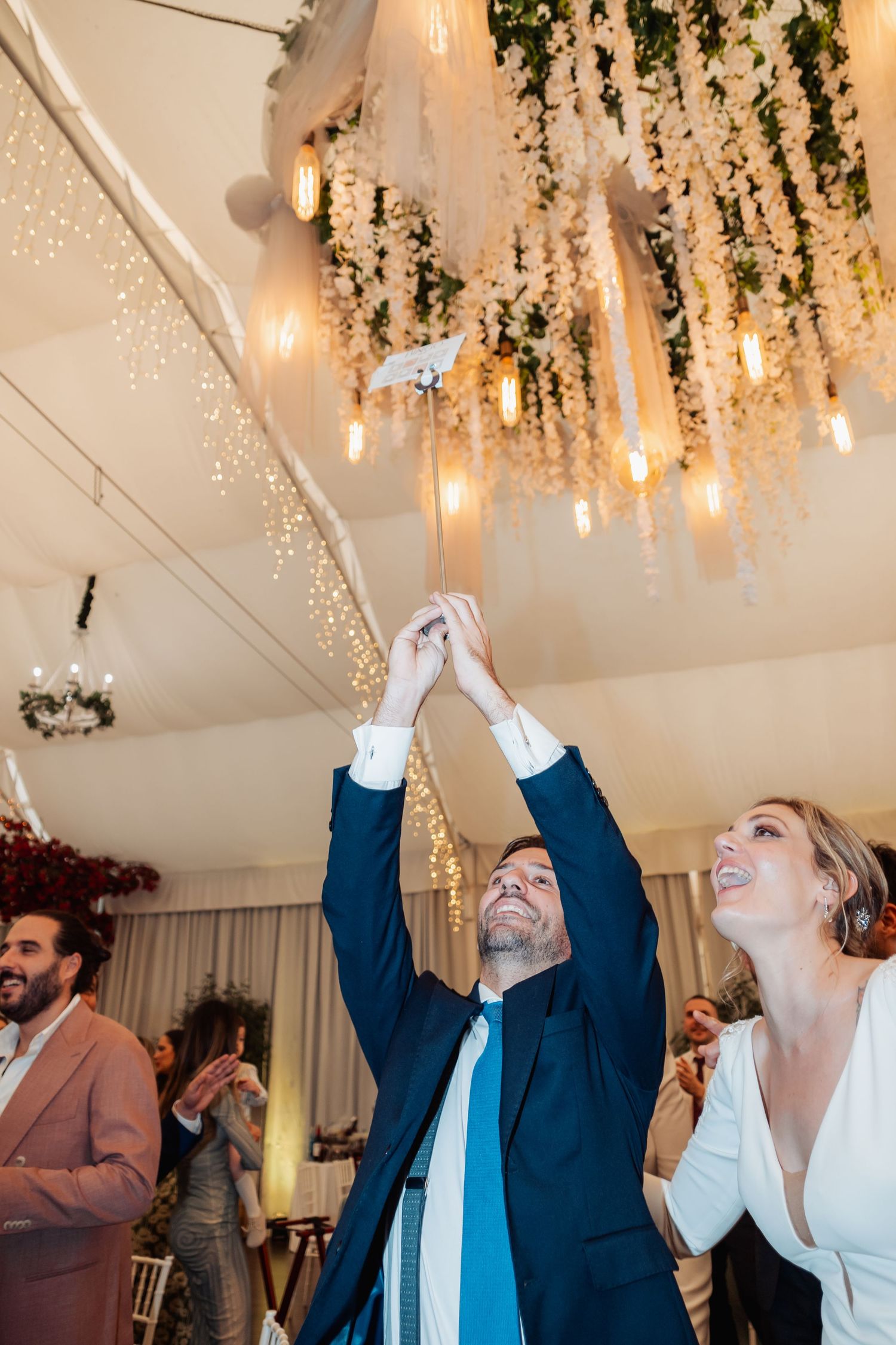 Groom releases white dove at wedding reception under floral chandelier with fairy lights.
