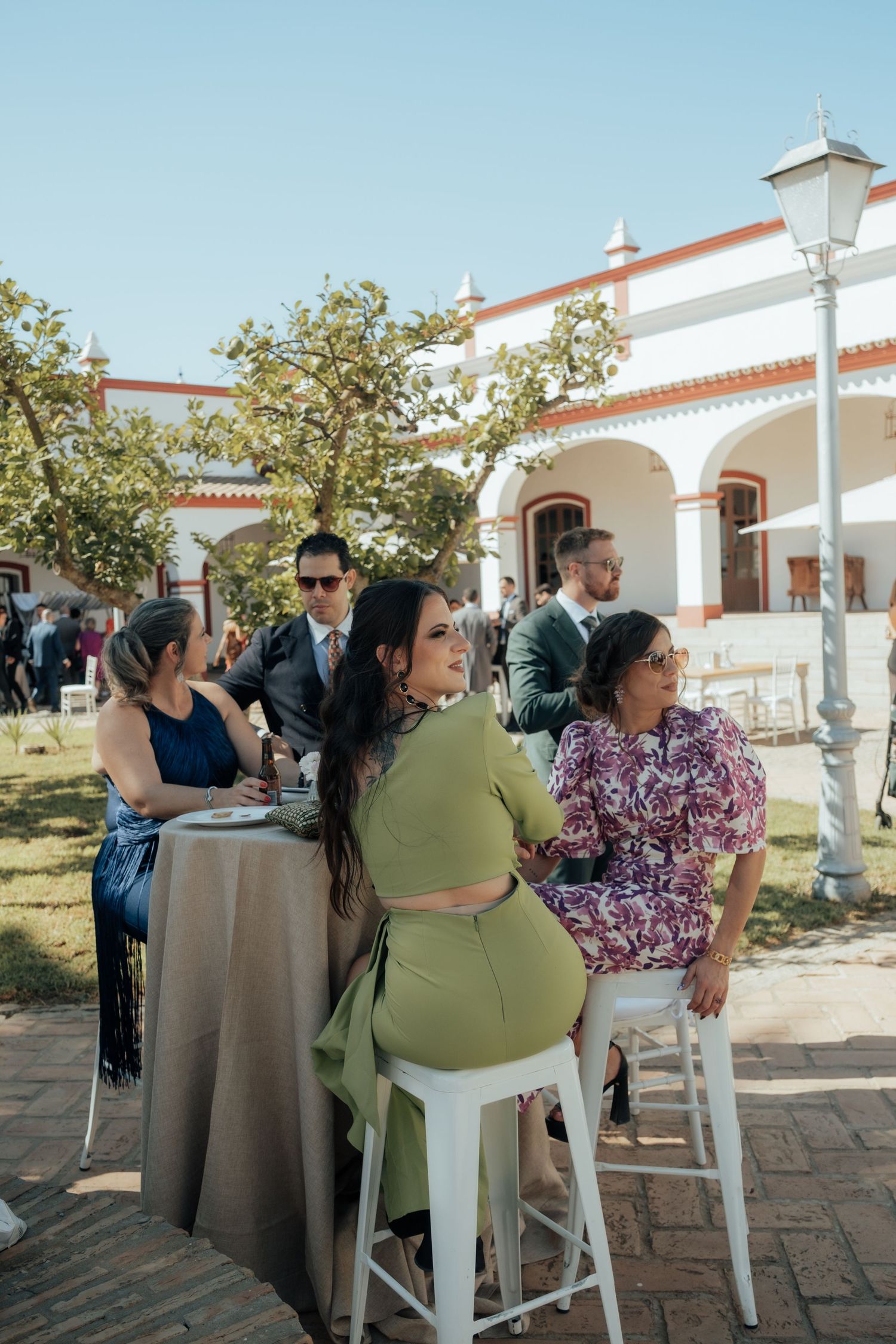 Wedding guests seated at outdoor high-top tables beside white Spanish-style venue.