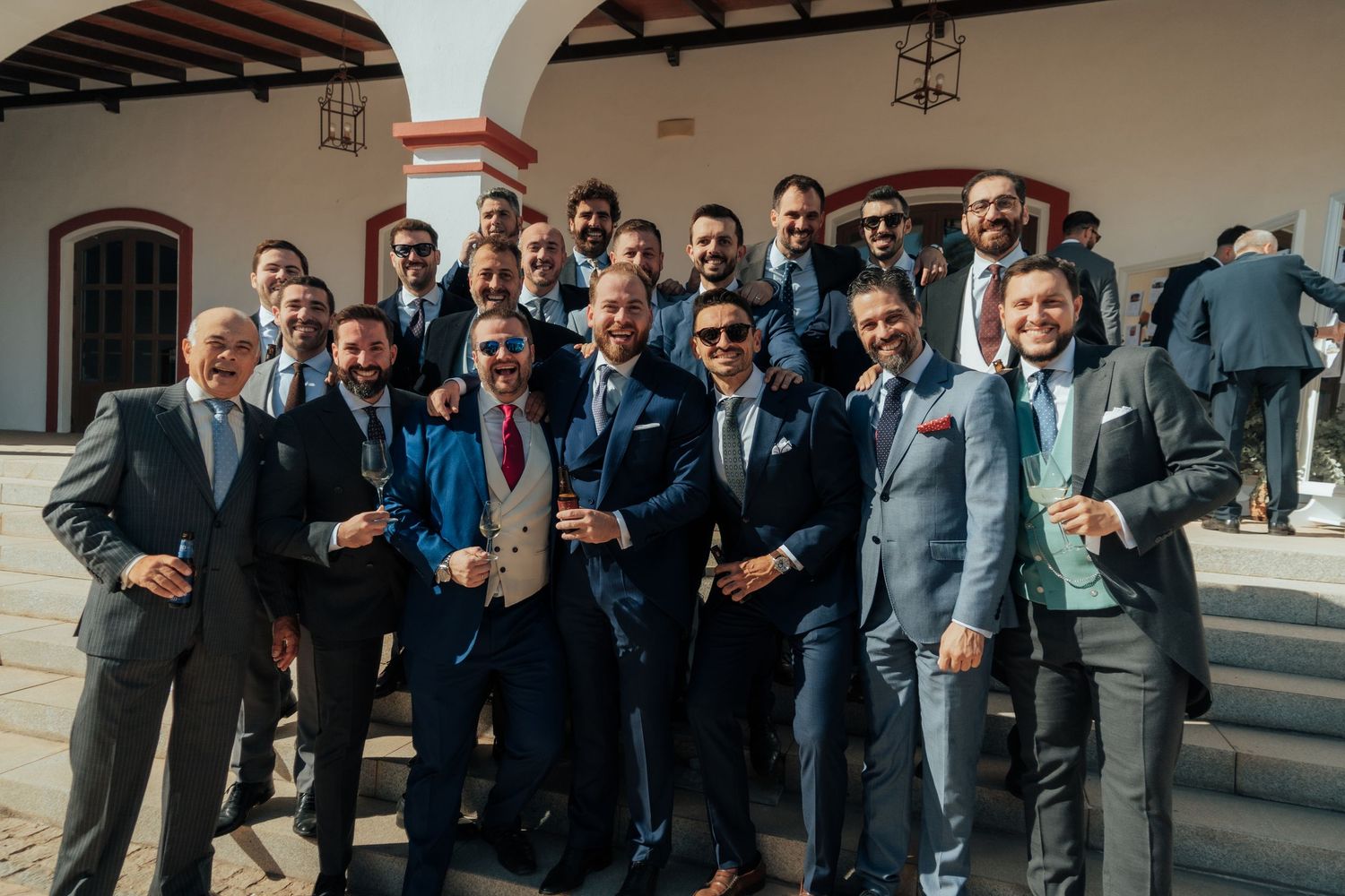 Large group of well-dressed men in suits smiling for a photo outside a white Spanish building.