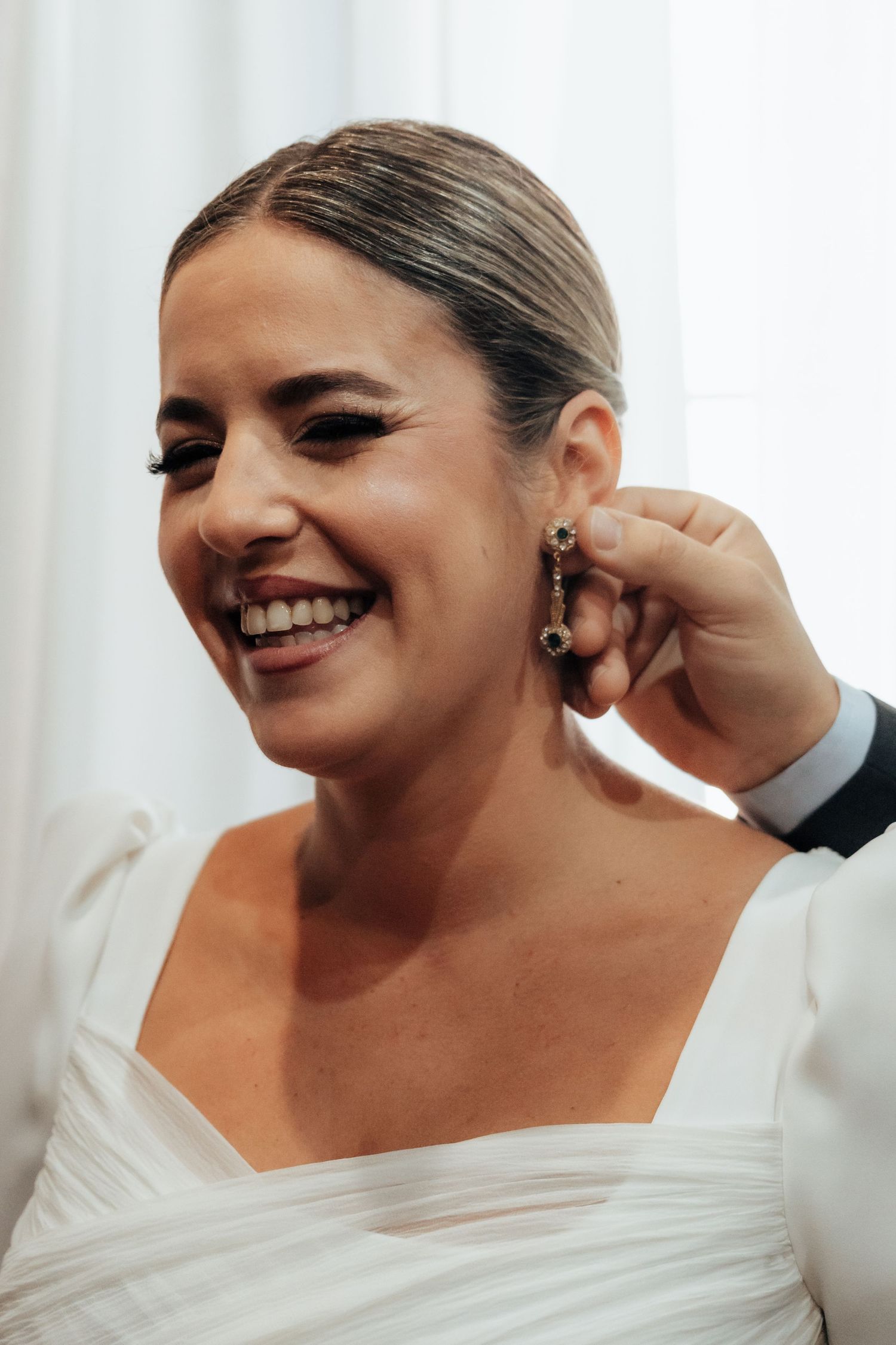 Smiling bride in white wedding gown having dangling earrings put on before her ceremony.