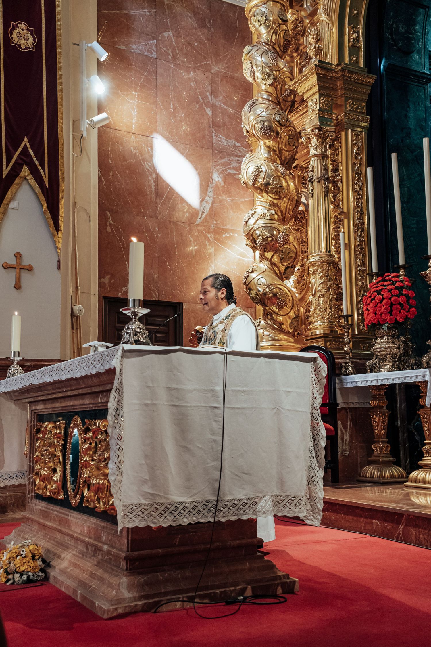 Catholic priest stands at ornate golden altar with candles and red flowers during wedding Mass ceremony.