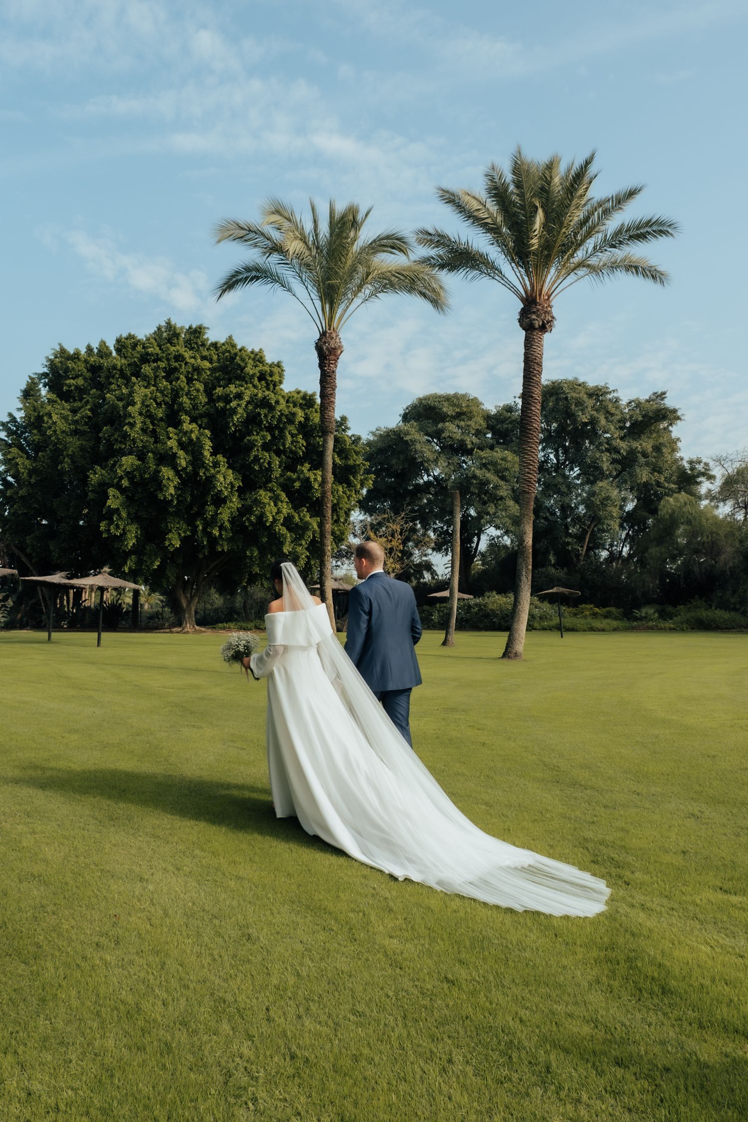 Bride and groom walking hand in hand across a lush green lawn with tall palm trees in background.