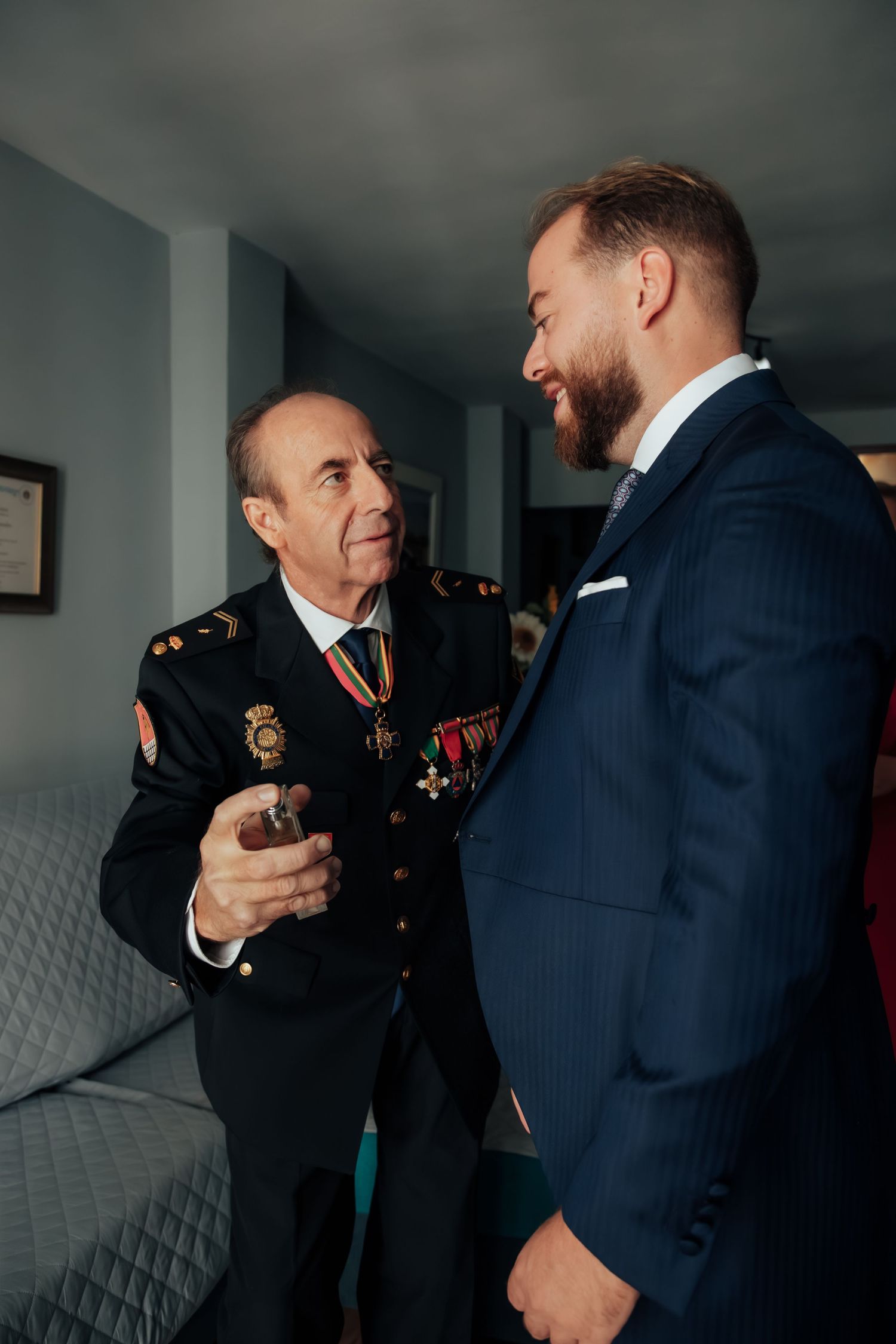 Groom in navy suit conversing with older man in decorated military uniform indoors.
