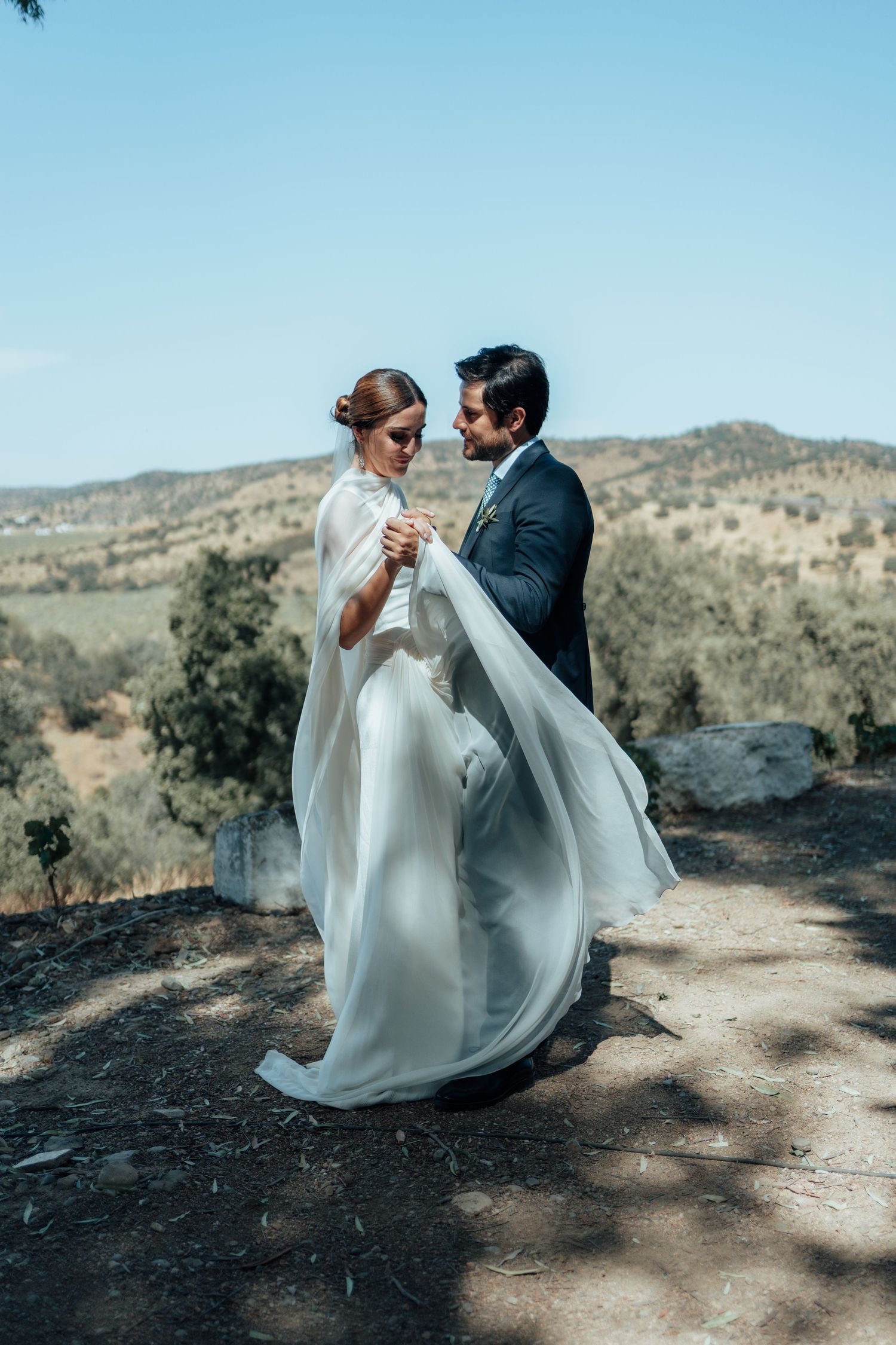 Bride in flowing white cape gown and groom in navy suit dancing with scenic mountain landscape behind.