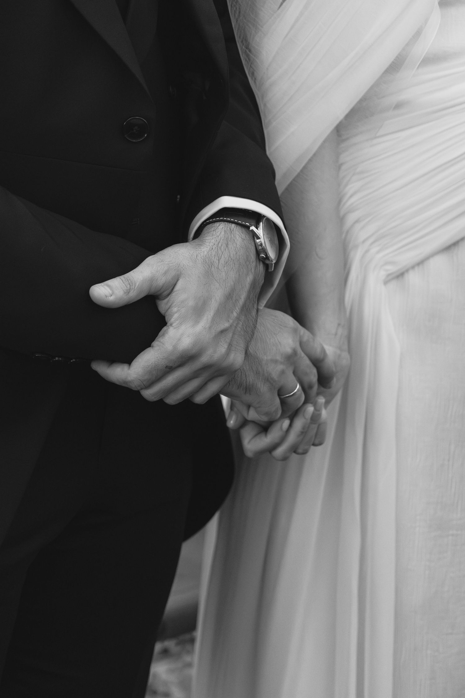 Black and white photo of bride and groom holding hands, wedding ring visible, at ceremony.