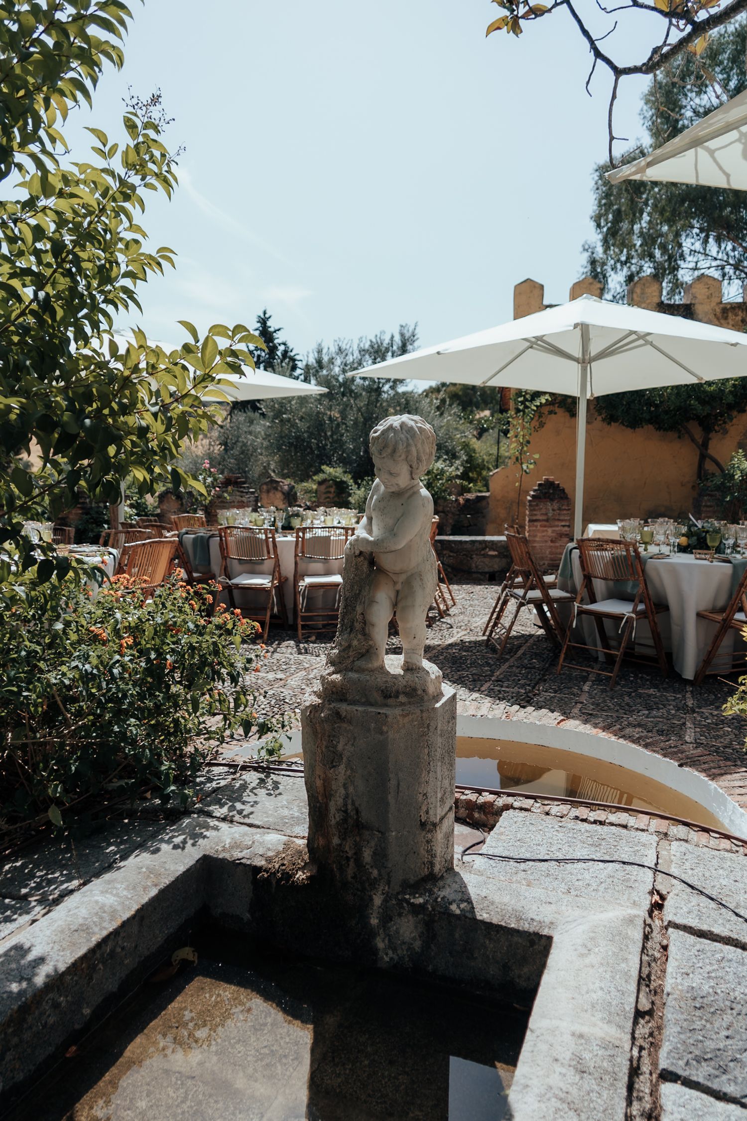 Stone cherub fountain in a sunlit garden wedding venue with white umbrellas and round tables in the background.