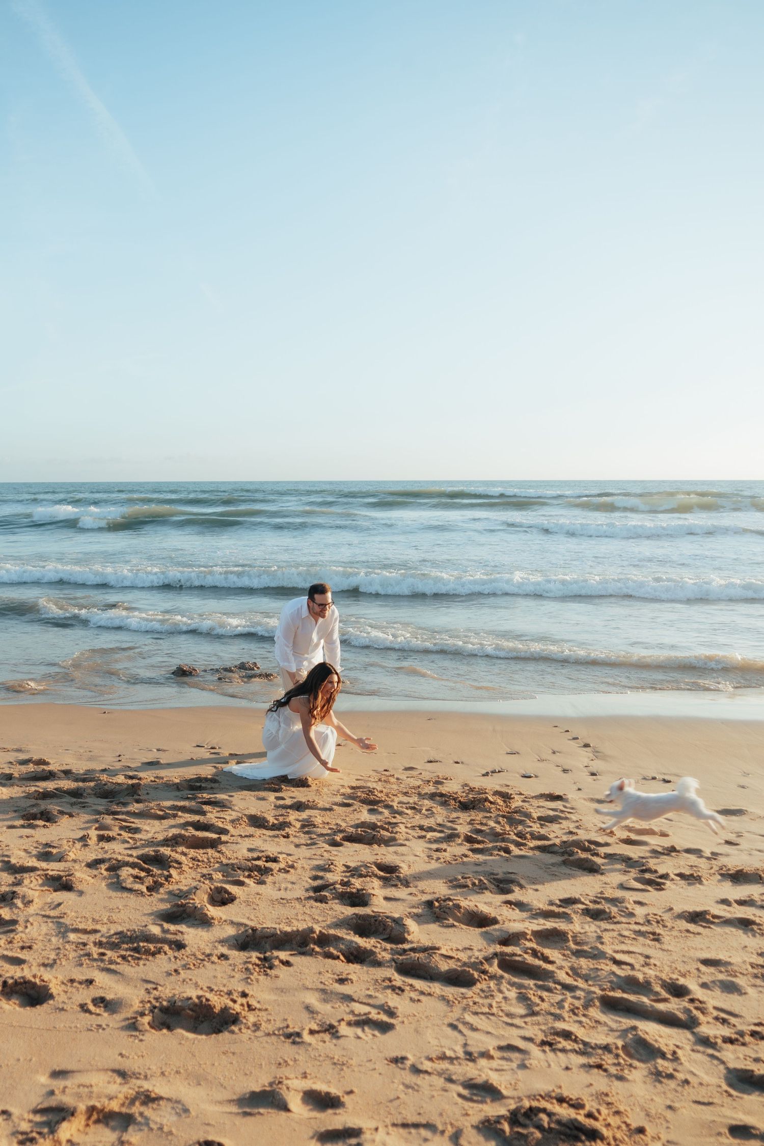 Couple in white attire walking romantically on a sunny sandy beach with ocean waves in the background.