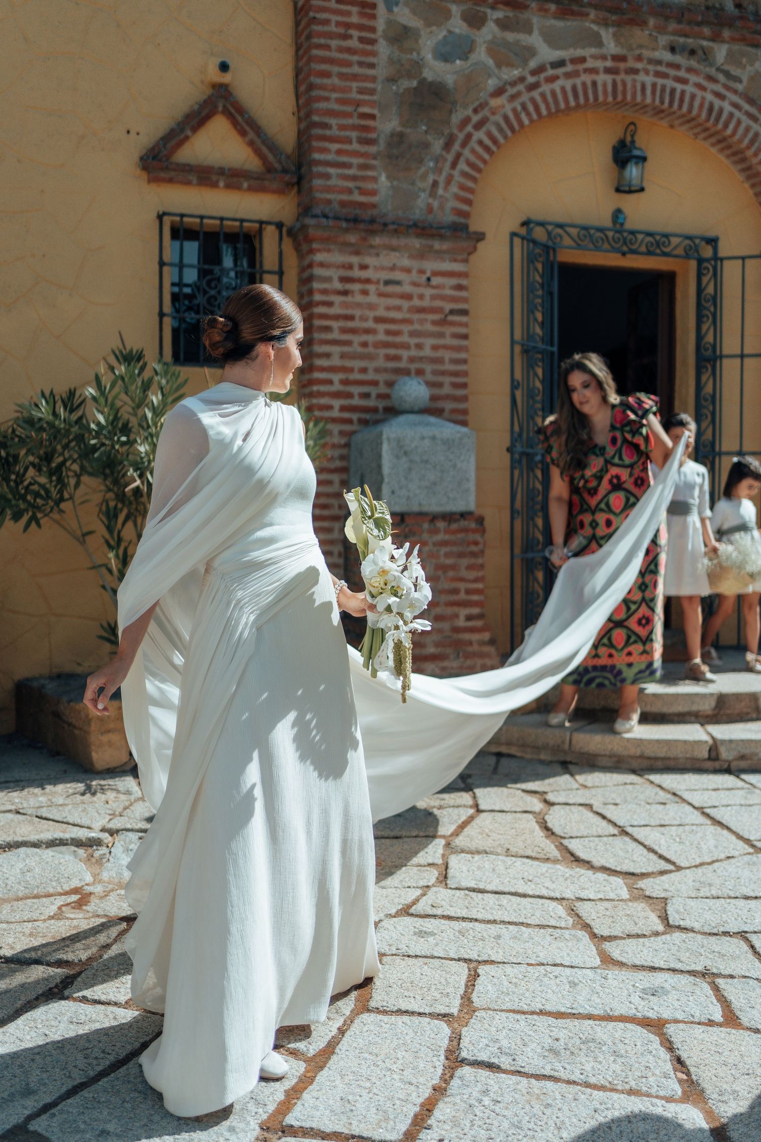 Bride in white flowing dress with train held by bridesmaid outside rustic brick church entrance.