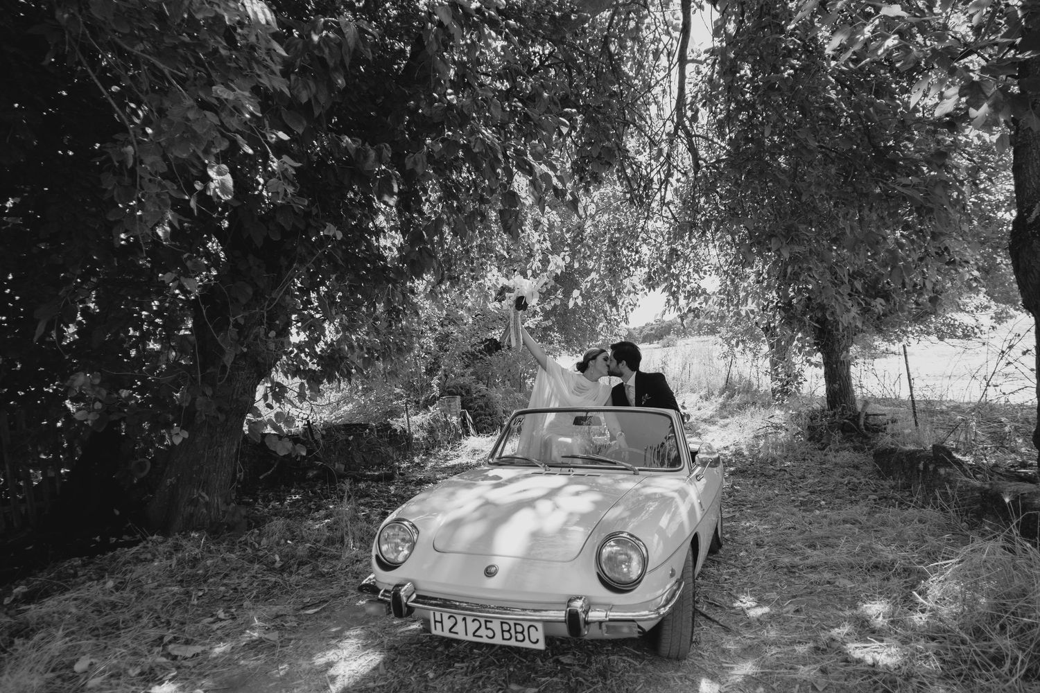 Black and white photo of newlyweds kissing in a white vintage convertible under shady trees.