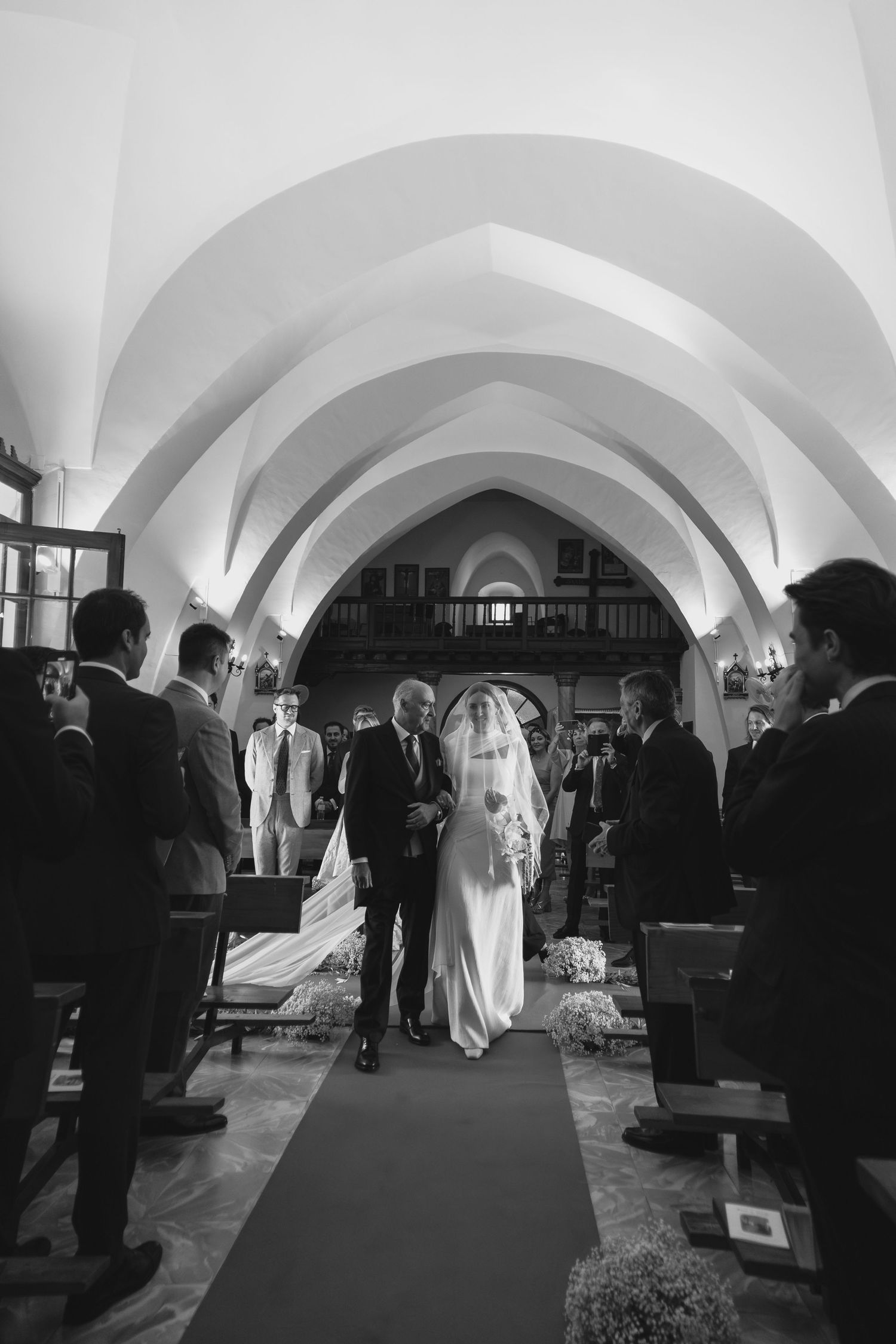 Black and white photo of bride walking down church aisle with vaulted ceiling and guests.