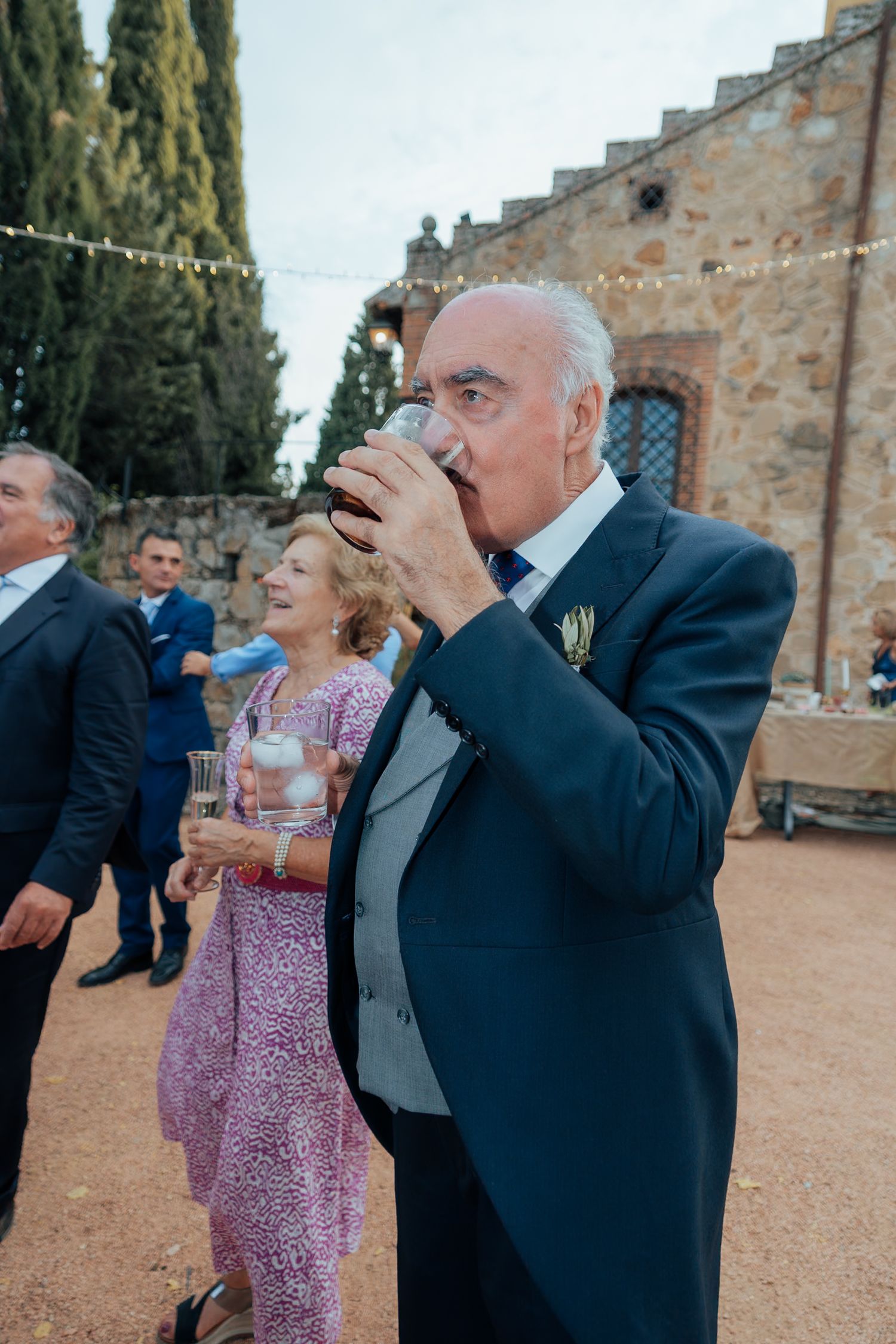 Elderly man in navy suit drinking from a glass at an outdoor wedding reception near a stone building.